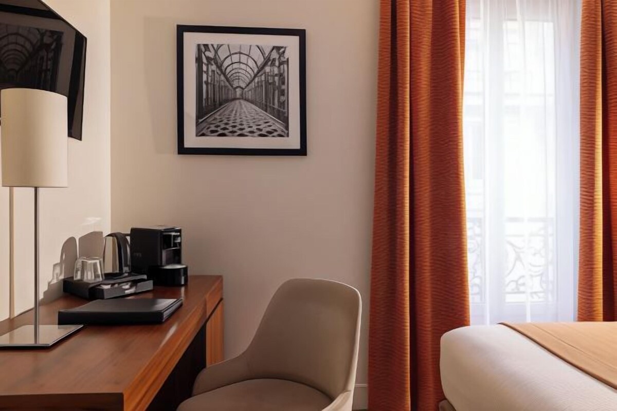 Hotelkamer met een bed, een bureau met lamp, een stoel, een ingelijst kunstwerk aan de muur en een raam met gordijnen.