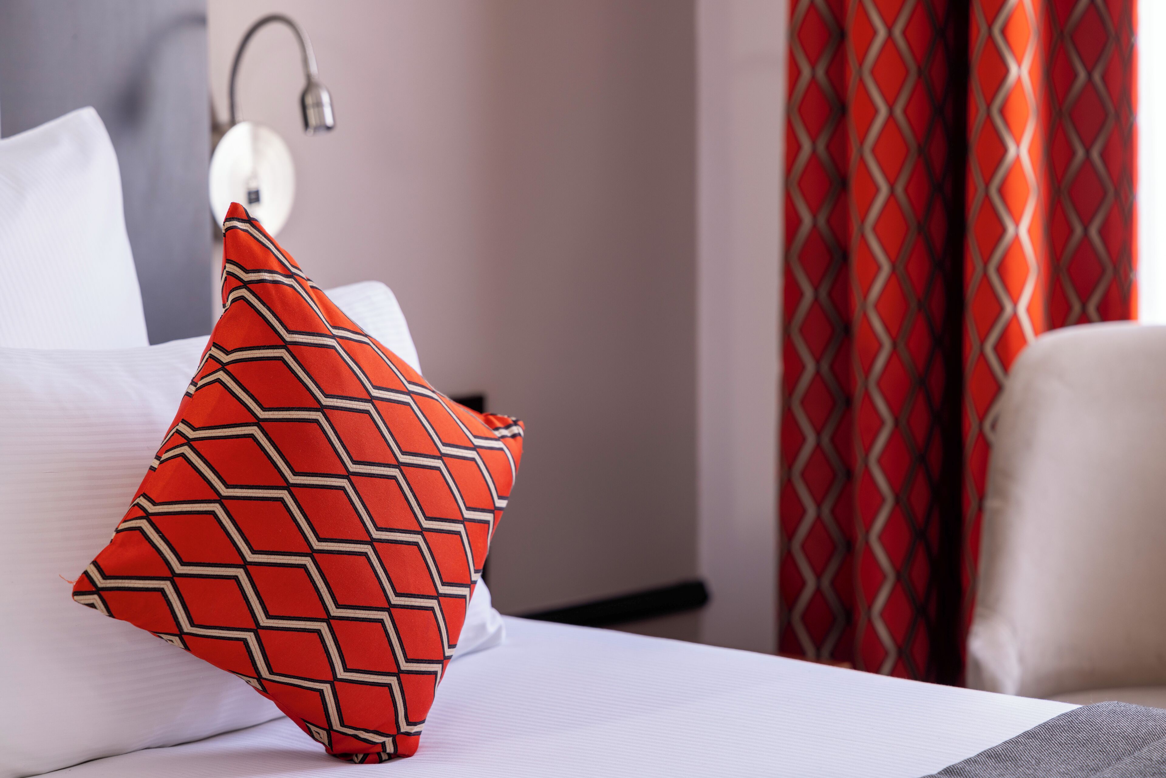 Une chambre d’hôtel avec un lit bien fait, un coussin rouge à motifs, un coussin blanc, une lampe fixée au mur et un rideau rouge.