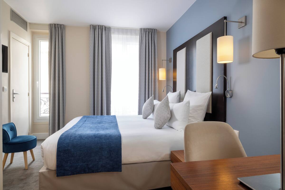 A hotel room with a bed, a desk, a chair, a TV, and a window with curtains.