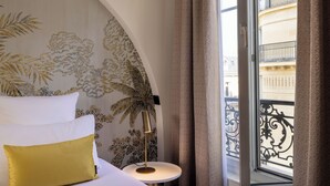 A hotel room with a bed, a round table with a lamp, a window with a view of a building, and a patterned wall. - Hôtel Elysées Bassano (Paris)