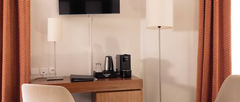 Habitación de hotel con televisor de pantalla plana montado en la pared, un escritorio con cafetera, una lámpara y dos sillas.