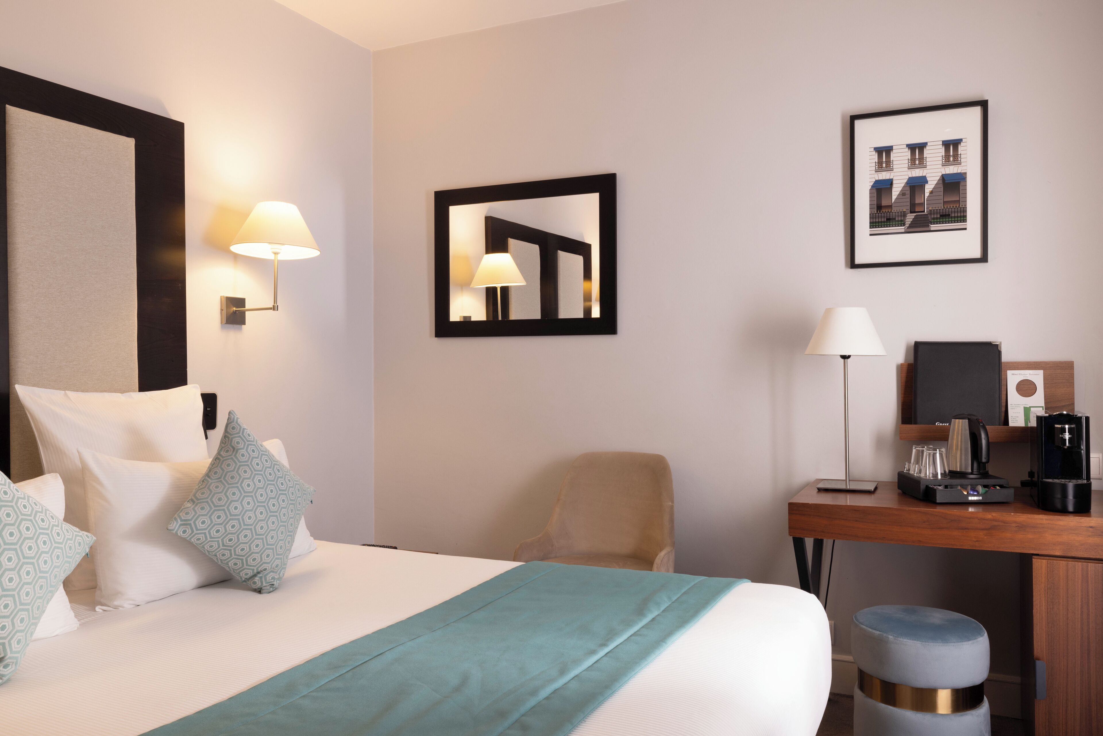 A hotel room with a bed, a desk, a chair, a coffee machine, and a framed picture on the wall.
