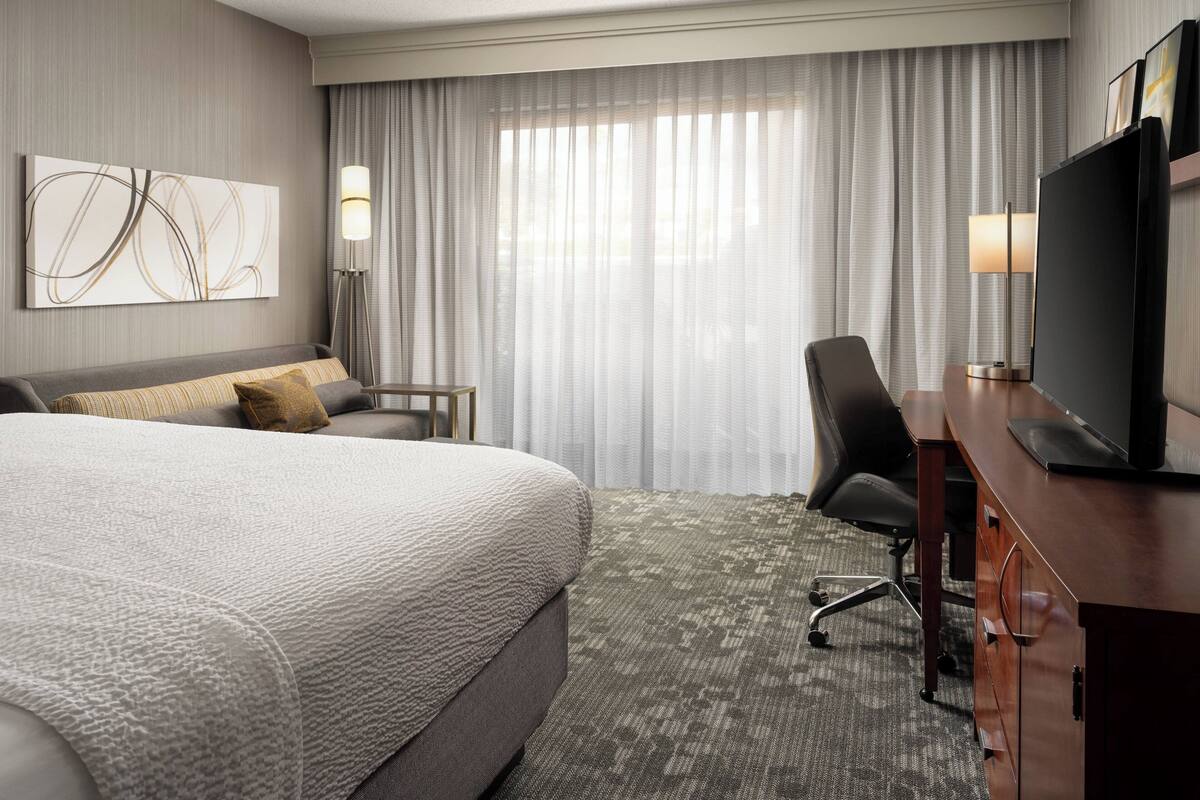 Habitación de hotel con una cama grande, un escritorio con televisor, una silla, una lámpara y un amplio ventanal con cortinas.