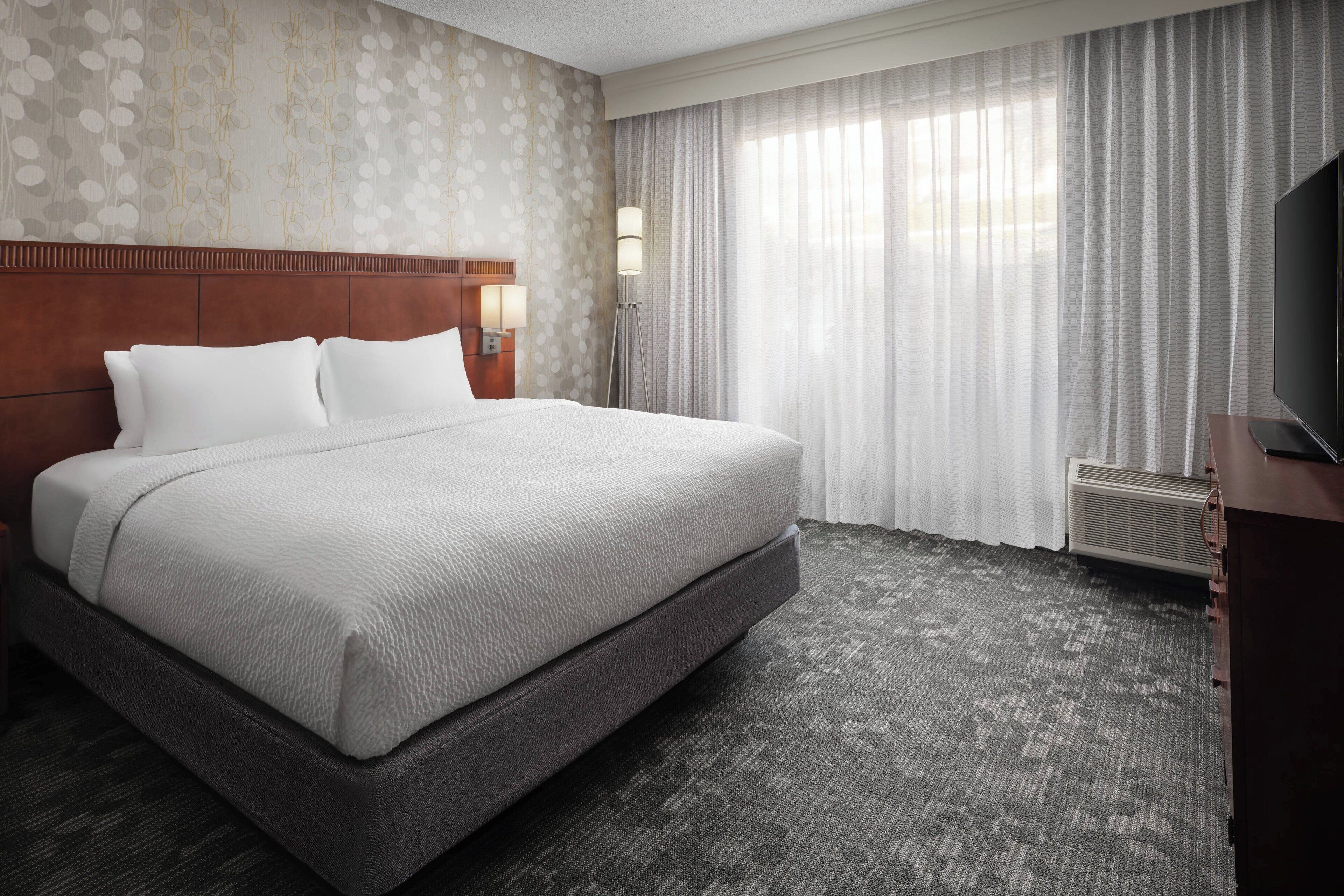 A hotel room with a large bed, a wooden headboard, a flat-screen TV, a nightstand with a lamp, and a window with sheer curtains.