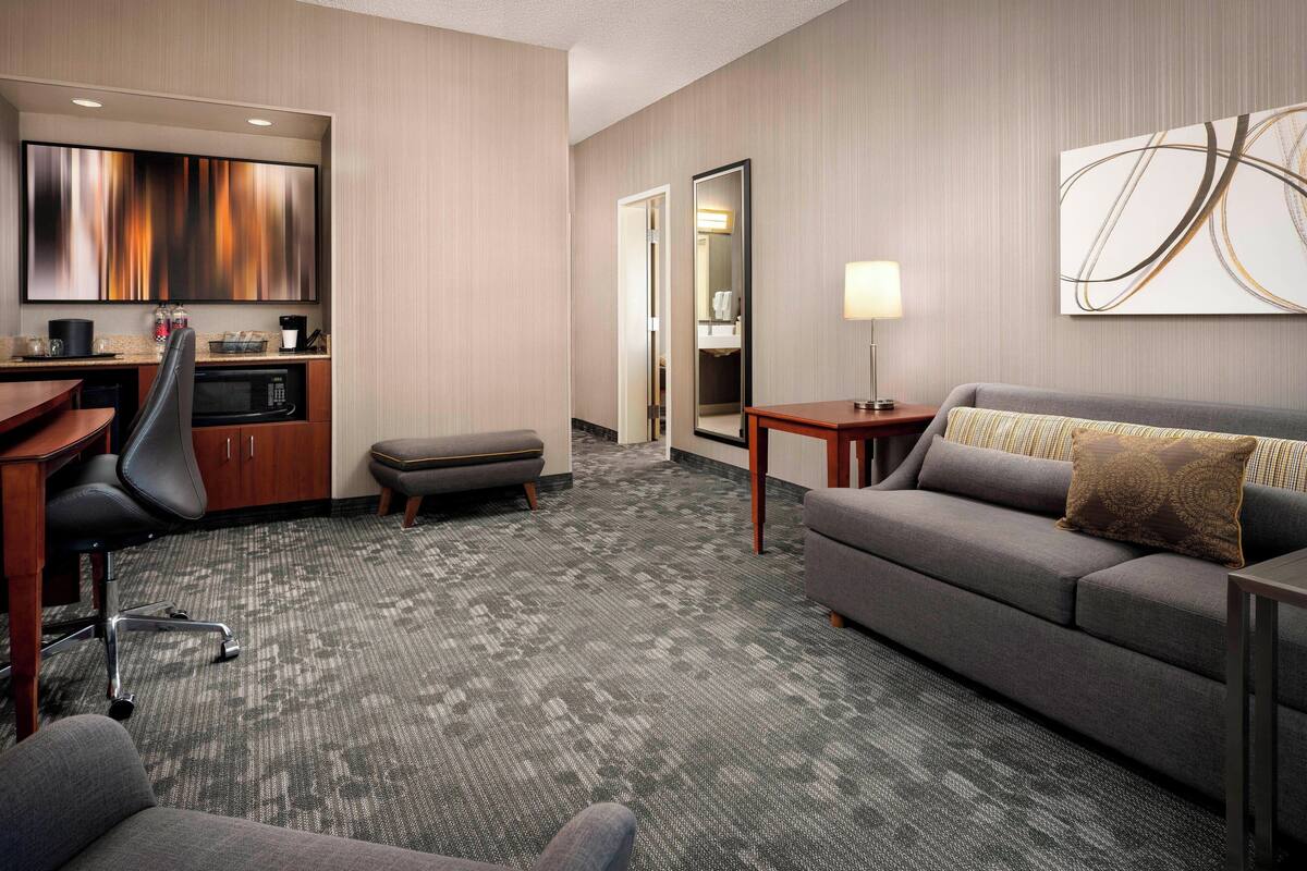 Habitación de hotel con un sofá gris, un escritorio con una computadora, una silla, un microondas y un cuadro abstracto grande en la pared.