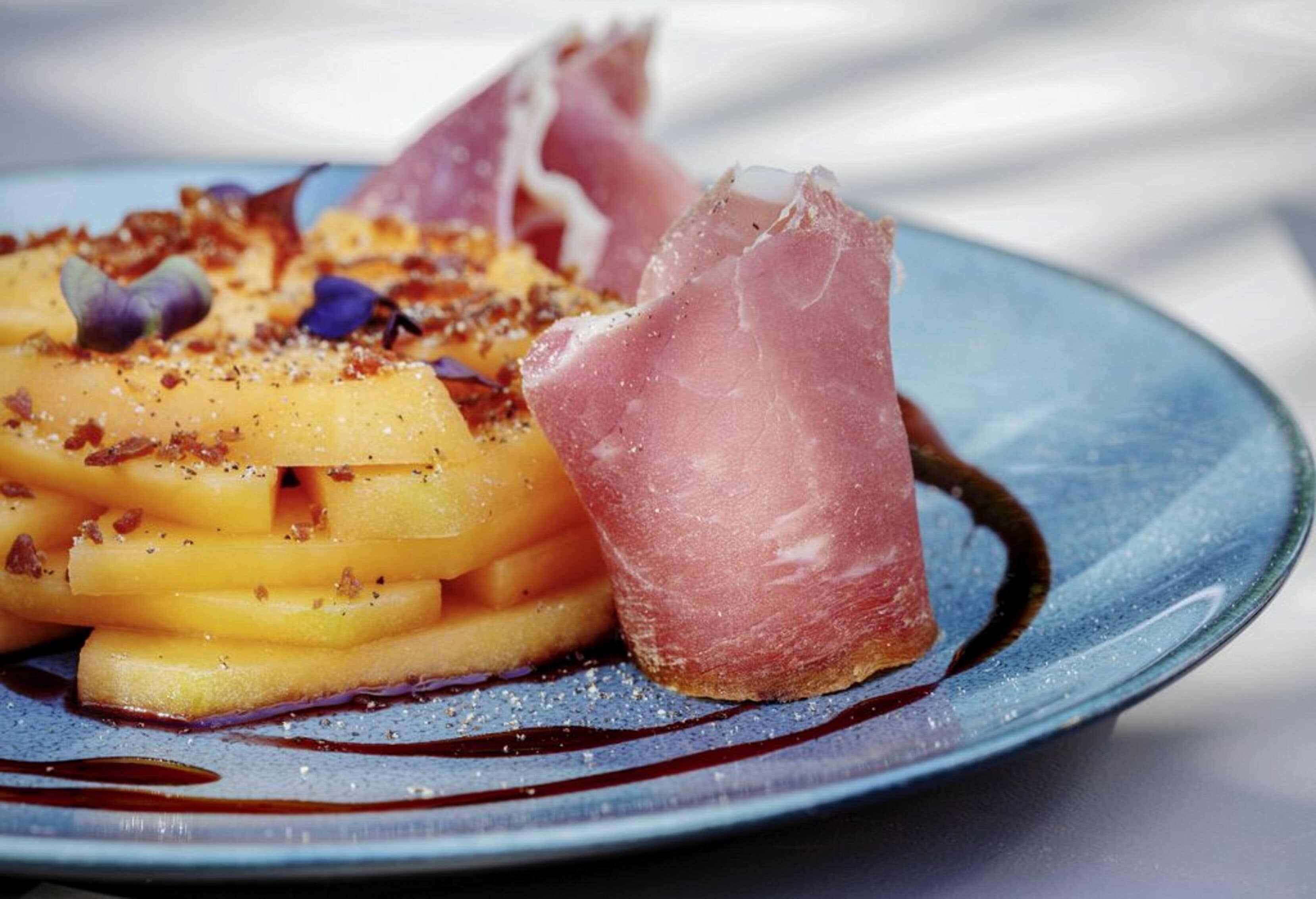 A plate with sliced fruit, possibly pears, topped with a pinkish slice of meat, possibly prosciutto, and sprinkled with herbs and spices.