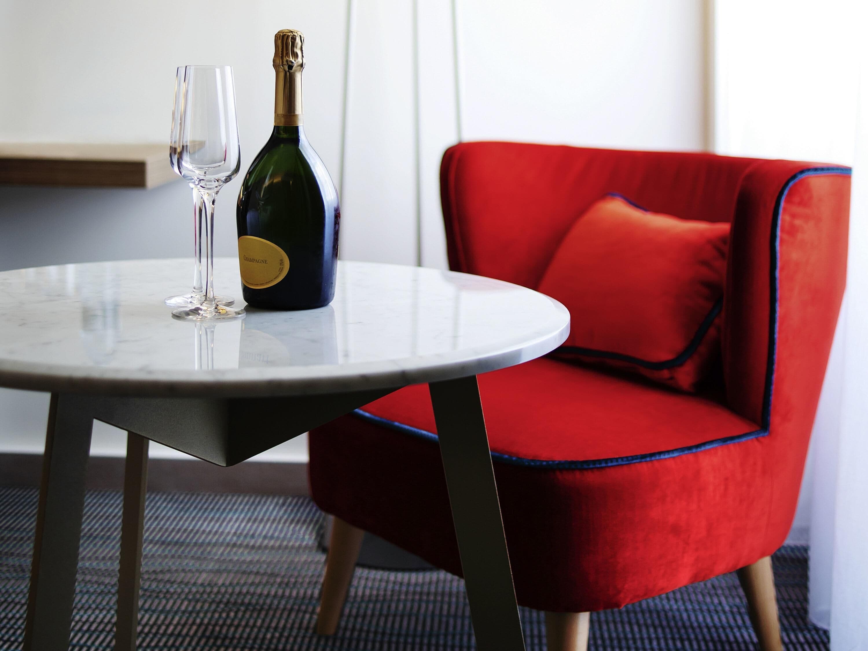Een ronde tafel met een champagnefles en een glas, een rode fauteuil met blauwe accenten, en een houten vloer.