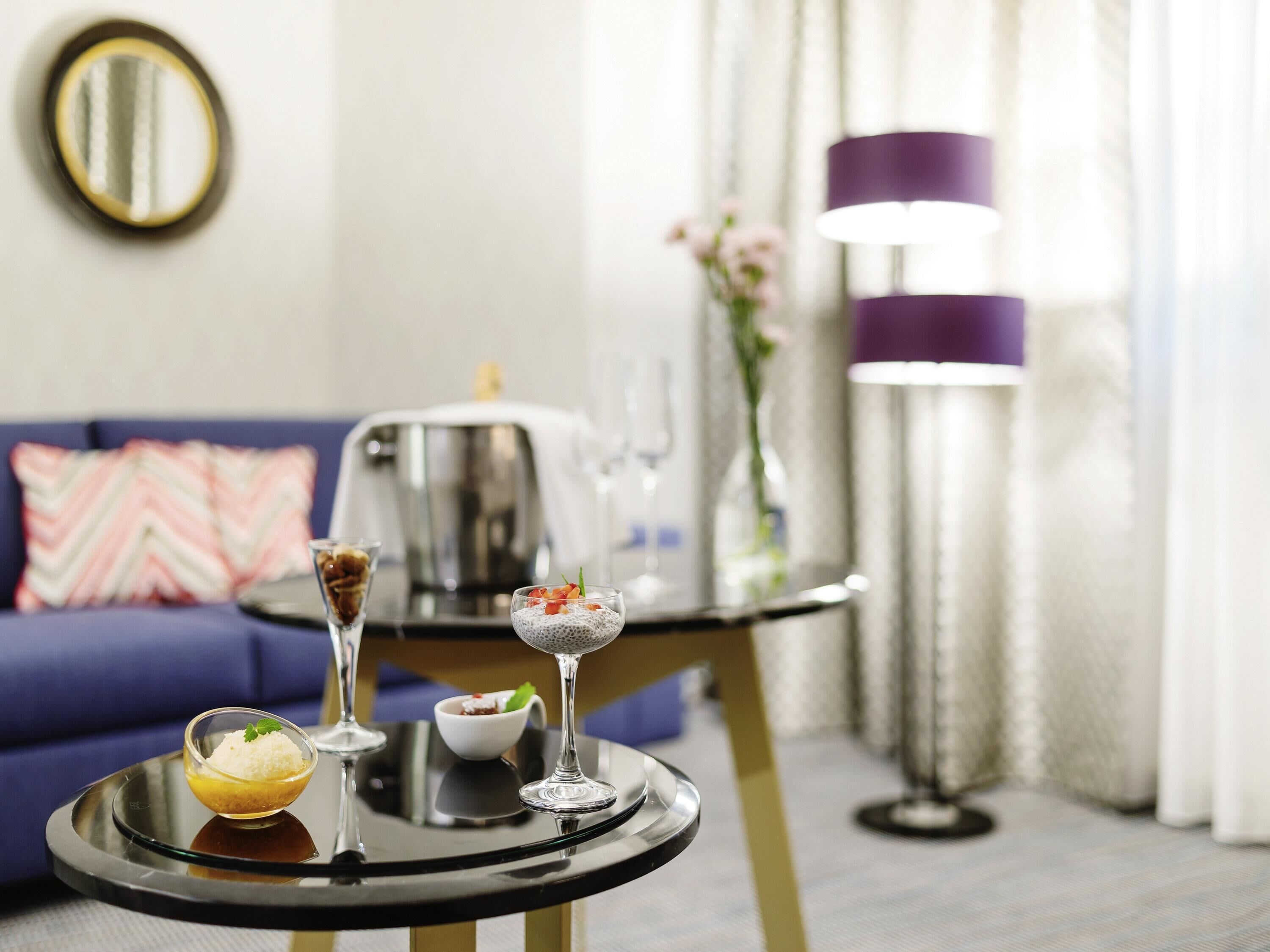 A dining table with a dessert and a glass of beverage, a sofa with a patterned cushion, a coffee maker, and a floor lamp.