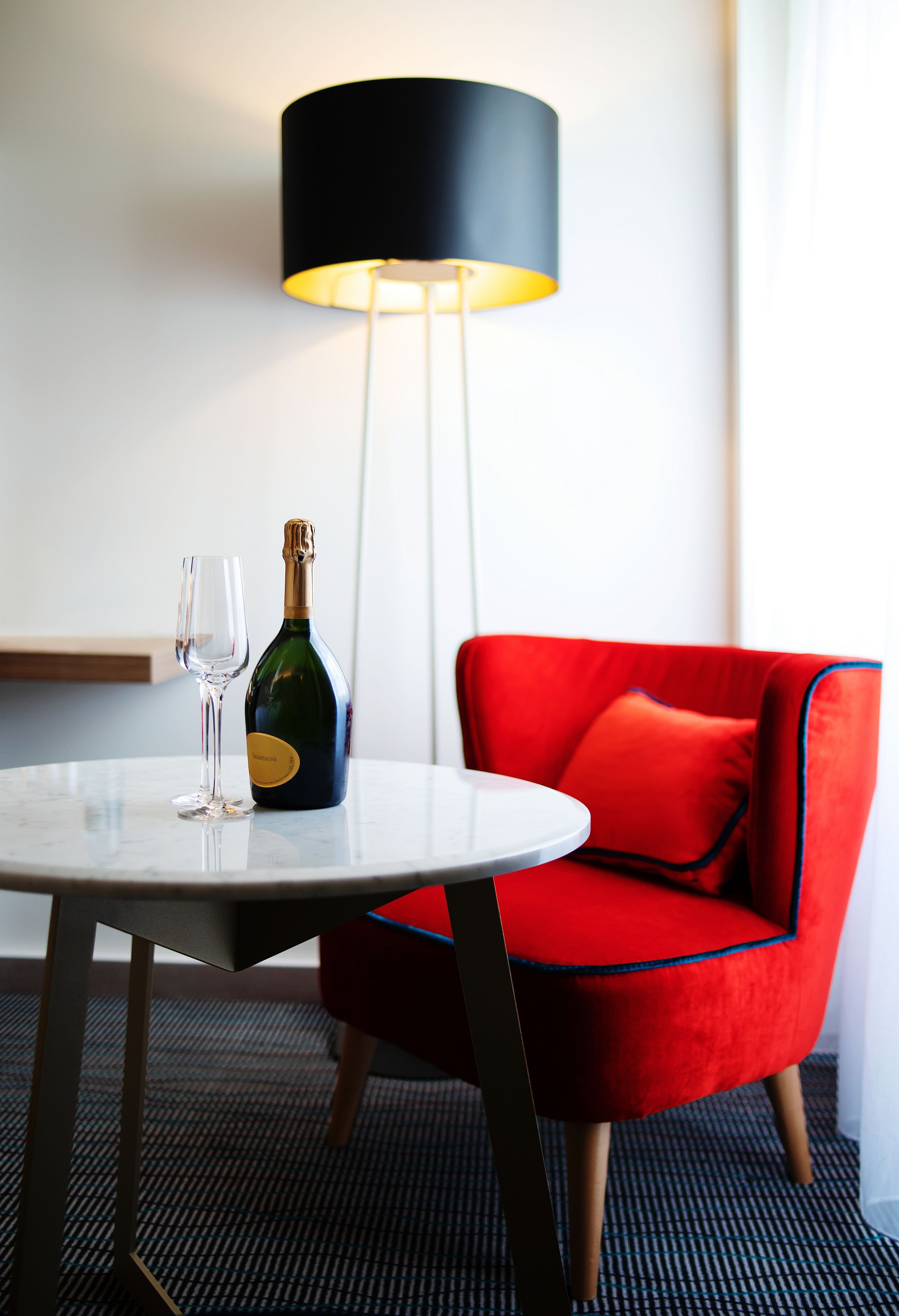 Et værelse med et rundt bord, en flaske champagne, et glas, en rød stol og en gulvlampe.