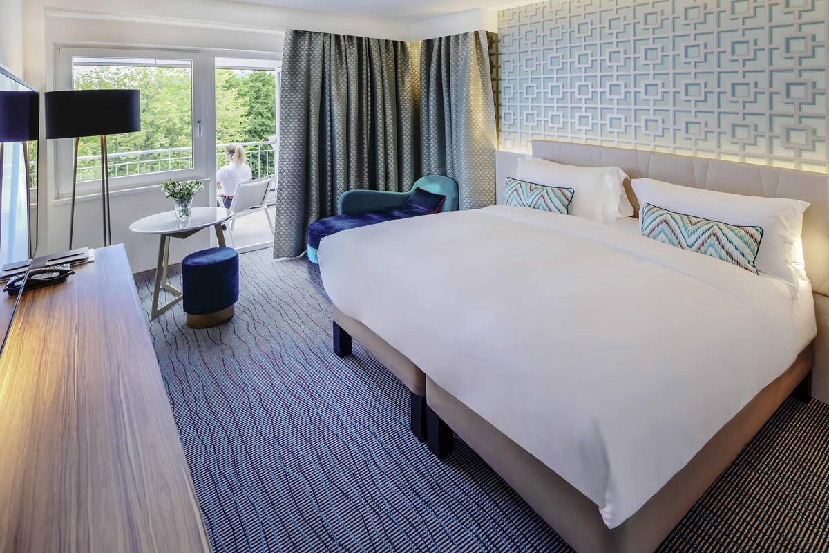 A hotel room with a large bed, a desk with a lamp, a small table with a chair, and a balcony with a view of trees.
