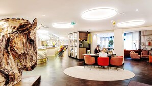 Un hall d’hôtel moderne avec une grande sculpture abstraite, un espace salon central doté de fauteuils orange et un bar avec des étagères.