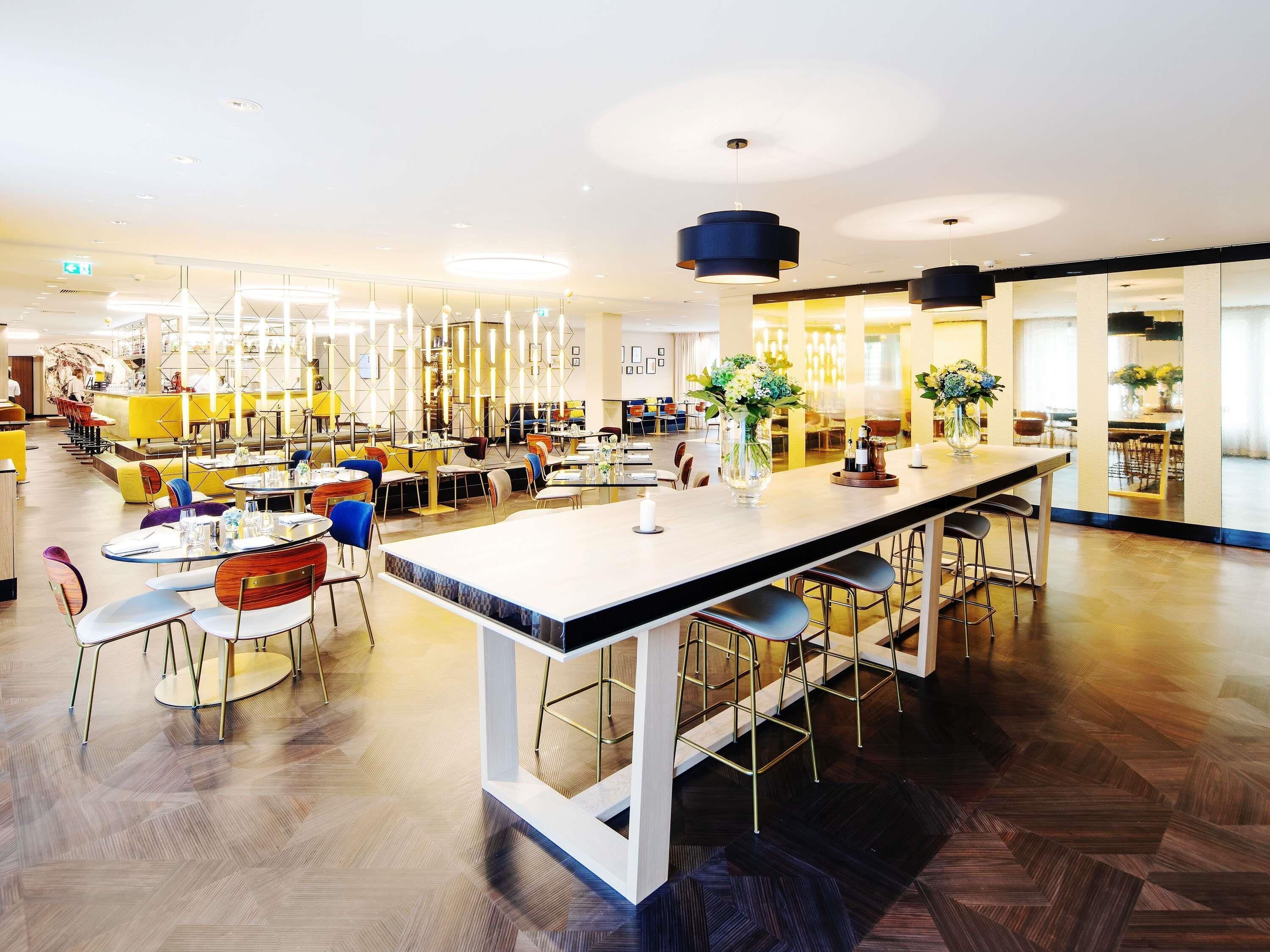 A modern restaurant with a long central bar, multiple seating areas, and contemporary lighting fixtures.