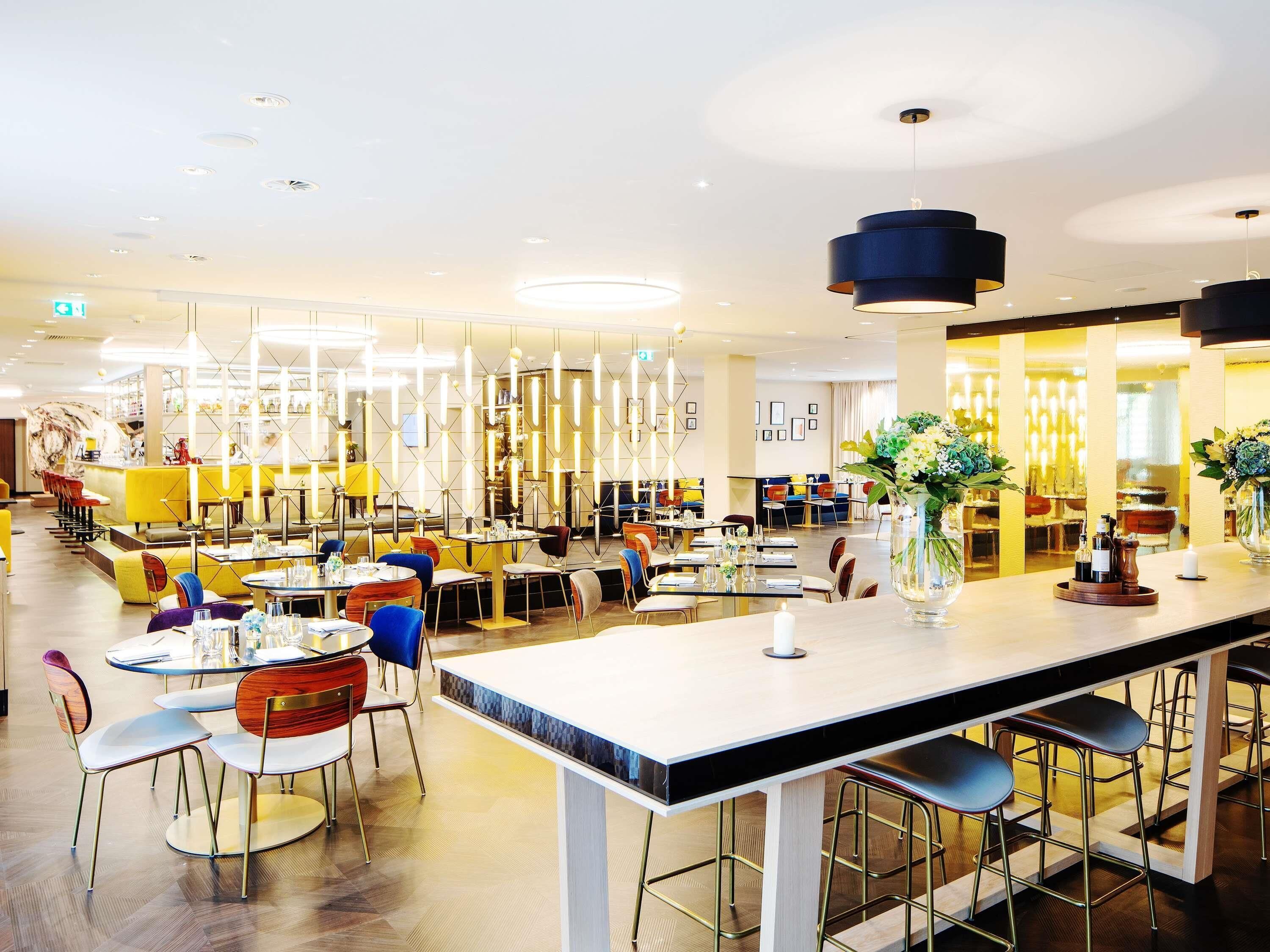 Ein modernes Restaurant mit hohen Decken, Pendelleuchten und verschiedenen Sitzmöglichkeiten, darunter Tische und Barhocker.