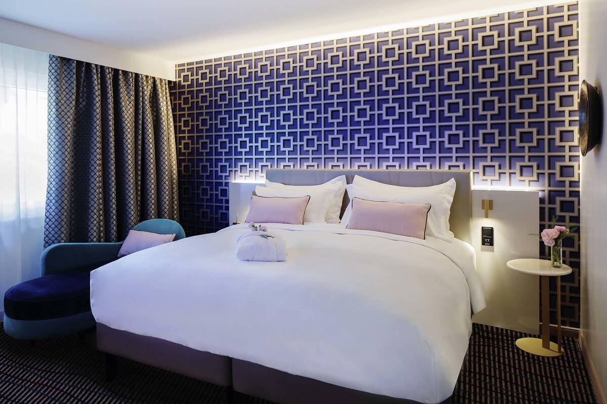 A modern hotel room with a large bed, a bedside table, a chair, and a patterned wall.