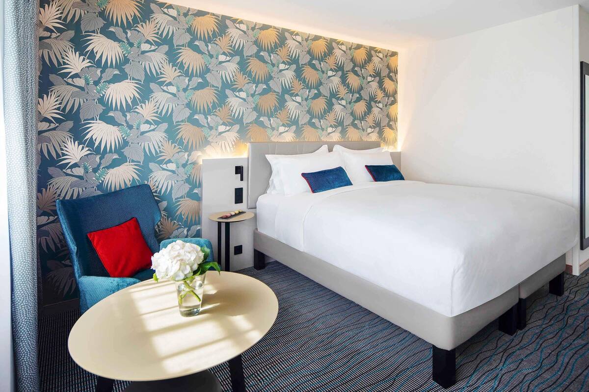A hotel room with a large bed, a small round table with a vase of flowers, a blue armchair, and a wall with a leaf pattern.