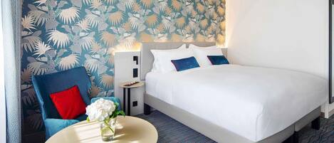 A hotel room with a large bed, a small round table with a vase of flowers, a blue armchair, and a wall with a leaf pattern.