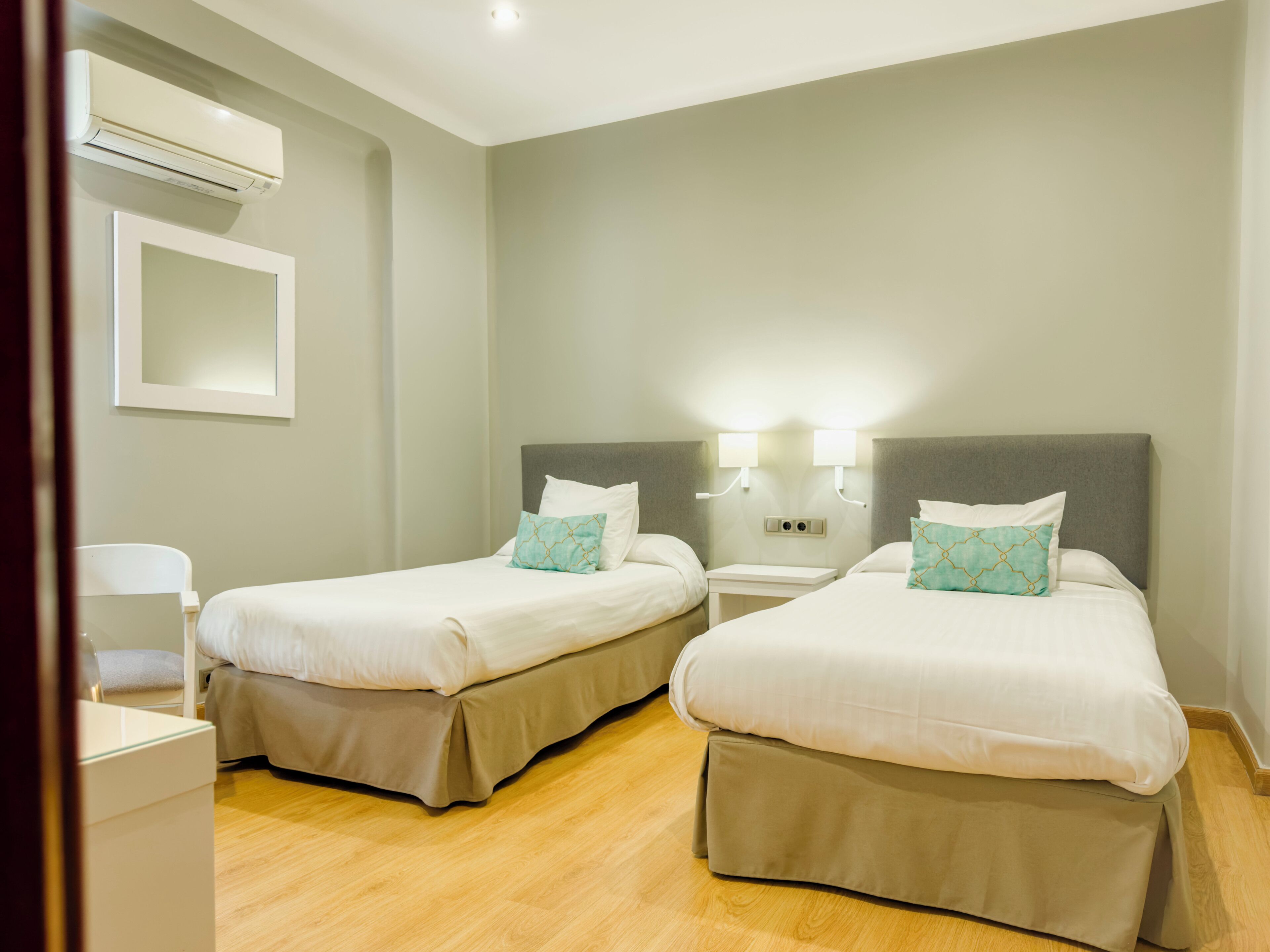 A hotel room with two beds, a bedside table, a nightstand, an air conditioning unit, and a mirror on the wall.