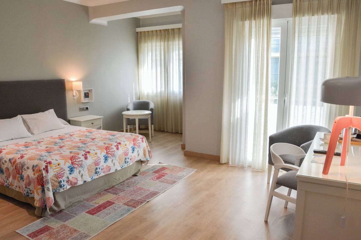 Habitación de hotel con cama, escritorio, silla, mesita y ventana con cortinas.