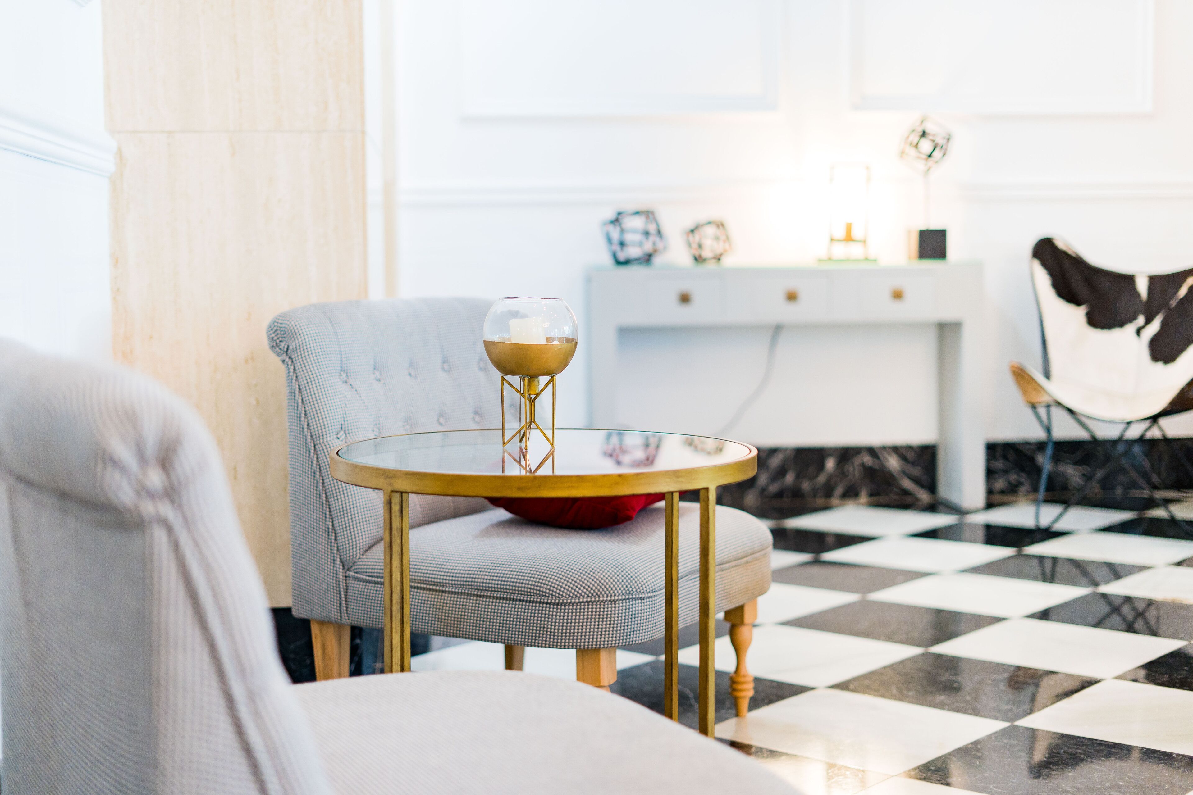 A room with a checkered floor, a round table with a glass top, a grey armchair, and a white console table with decorative items.