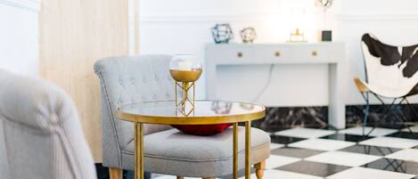 A room with a checkered floor, a round table with a glass top, a grey armchair, and a white console table with decorative items.