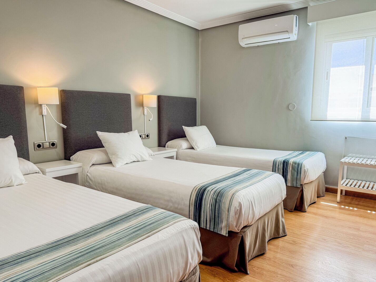 A hotel room with three beds, each with a pillow and a bedside table. There is a wall-mounted air conditioner and a window with blinds.