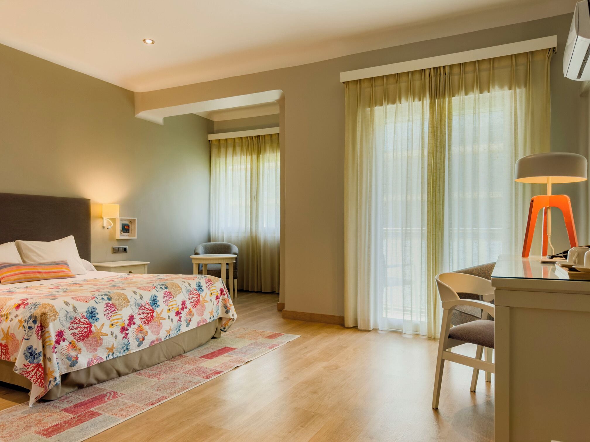 A hotel room with a bed, a desk, a chair, a lamp, and a window with curtains.