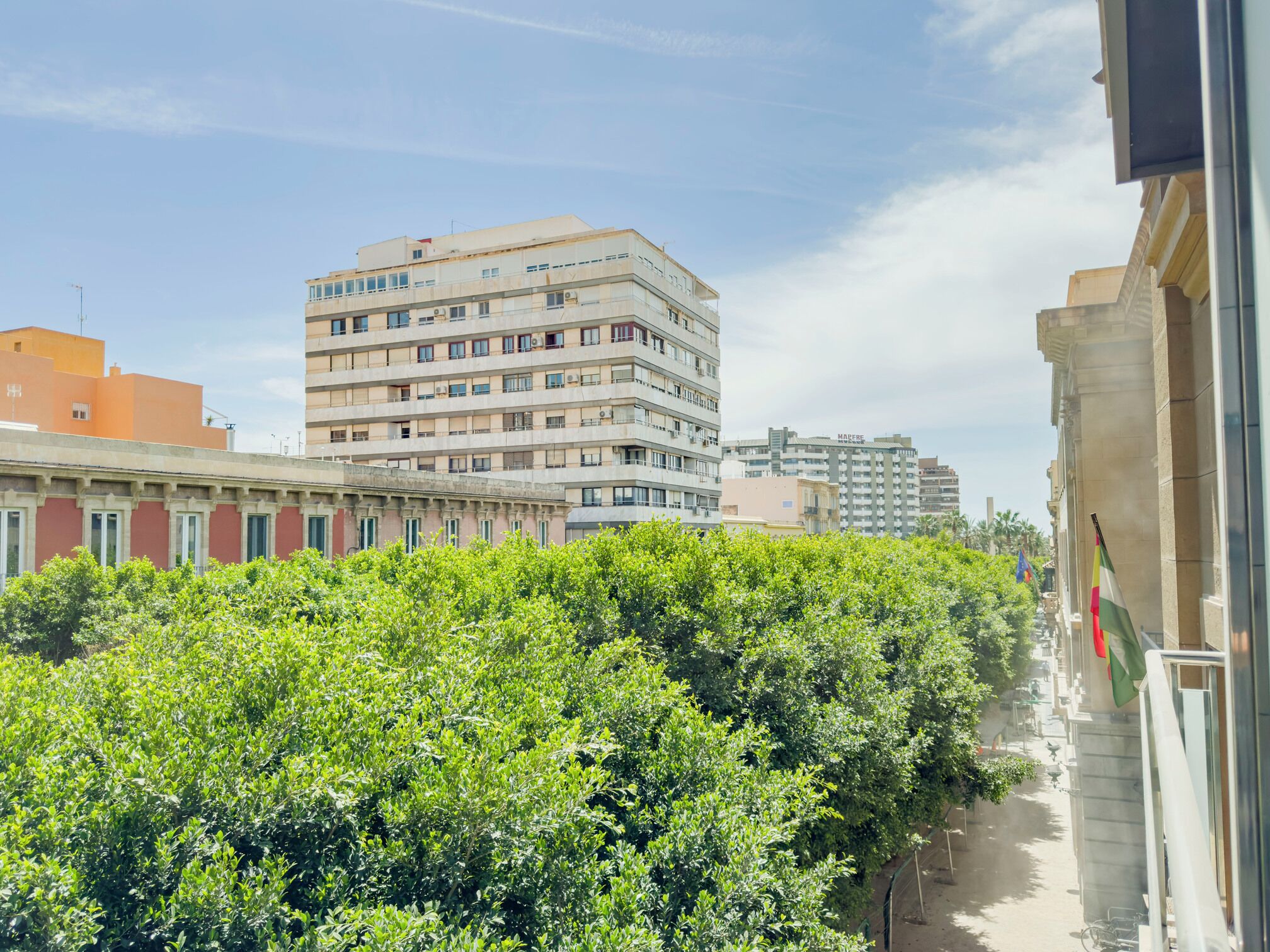 Un edificio de varios pisos con balcones, cielo despejado y un seto verde espeso al frente.