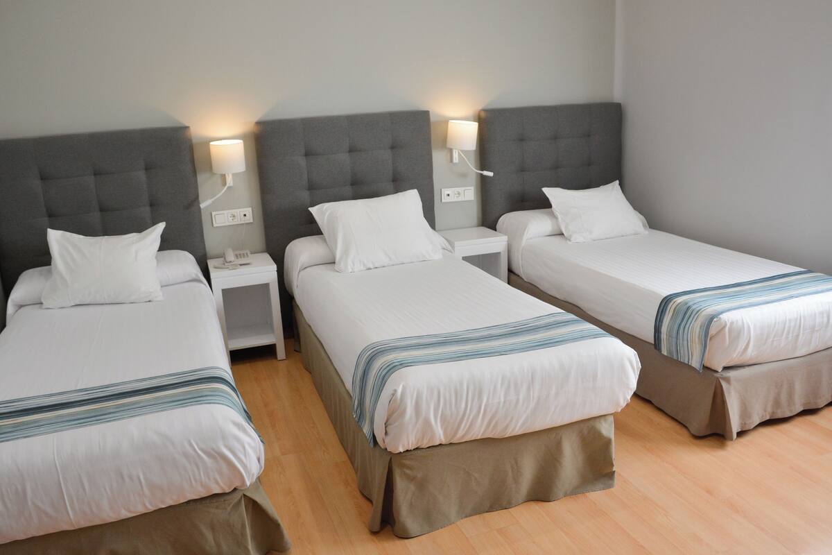 Habitación de hotel con tres camas individuales, cada una con cabecero gris y ropa de cama blanca. Hay mesitas de noche con lámparas y un enchufe.
