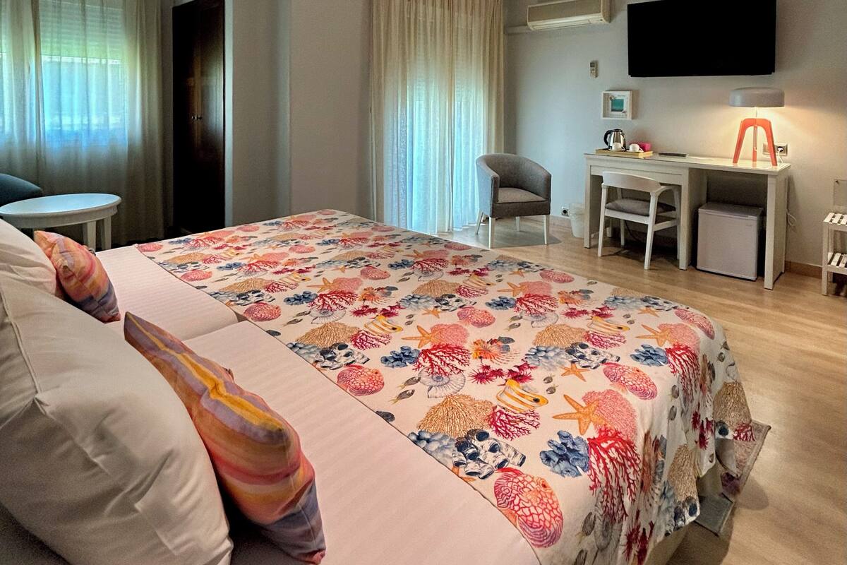 Habitación de hotel con cama, escritorio, silla, televisor y ventana con cortinas.