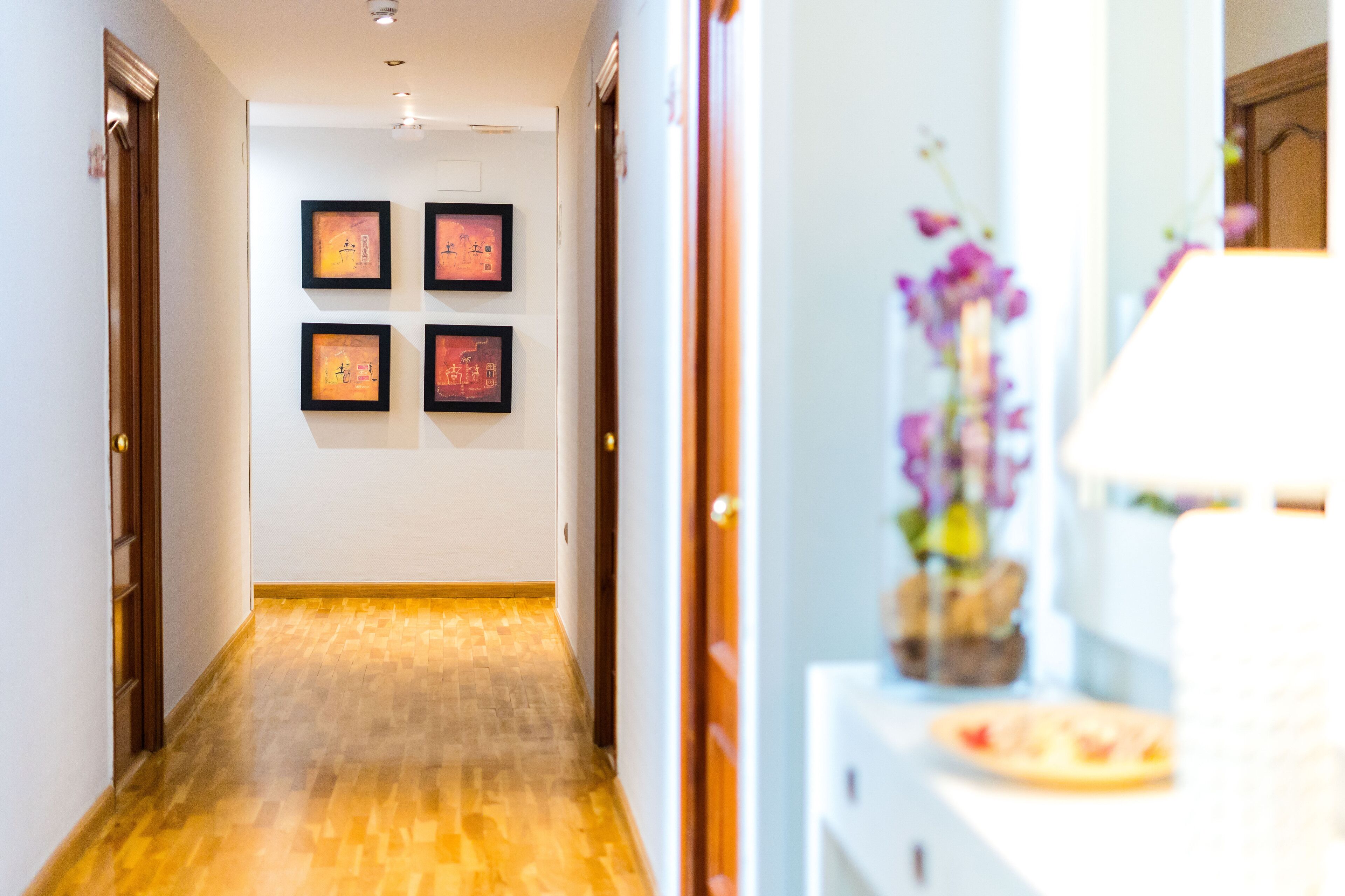 Un pasillo de hotel con piso de madera, obras de arte enmarcadas en las paredes y un jarrón decorativo con flores sobre una consola blanca.