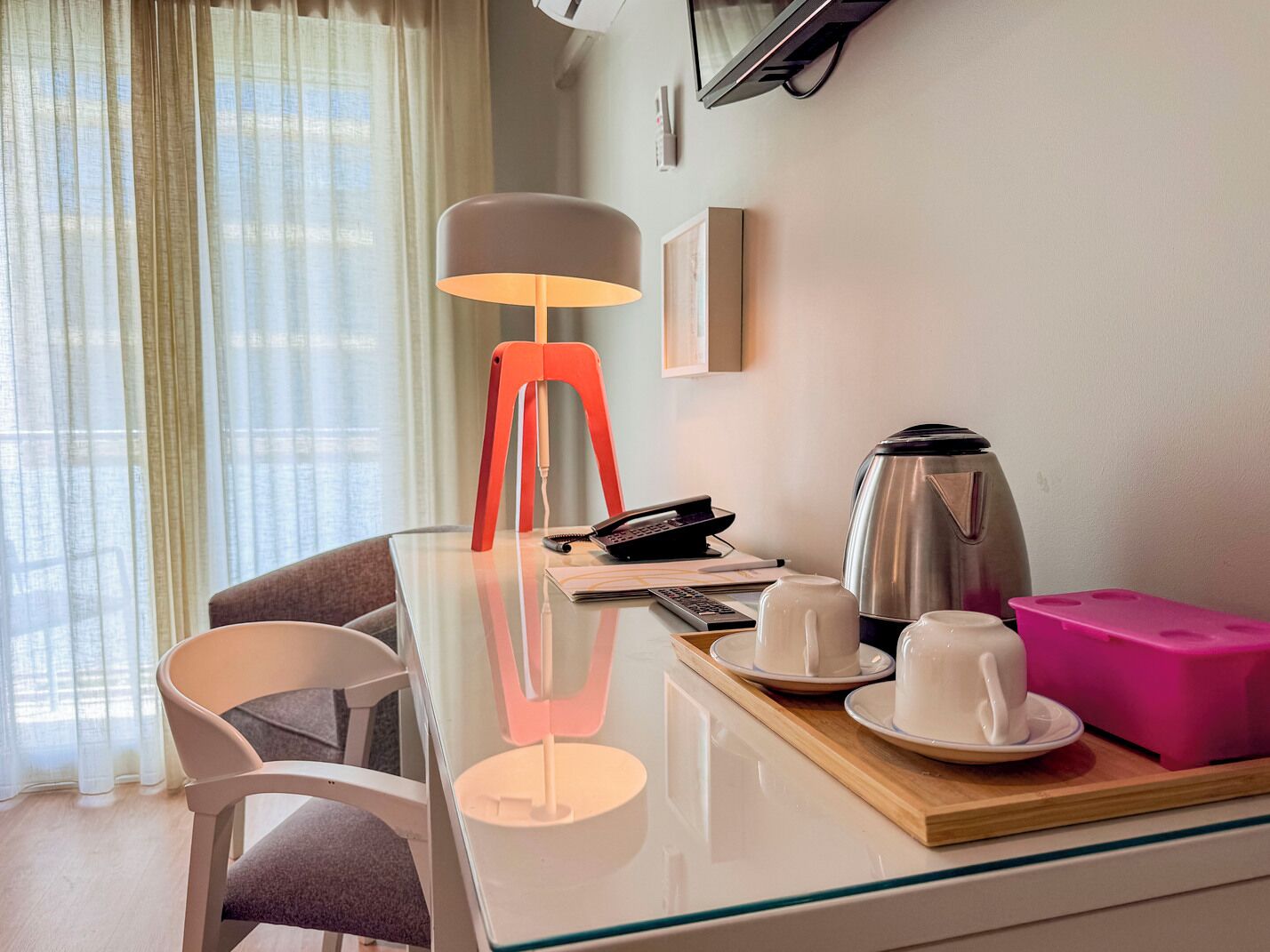 A hotel room with a glass table, a red lamp, a coffee maker, and a pink storage box.