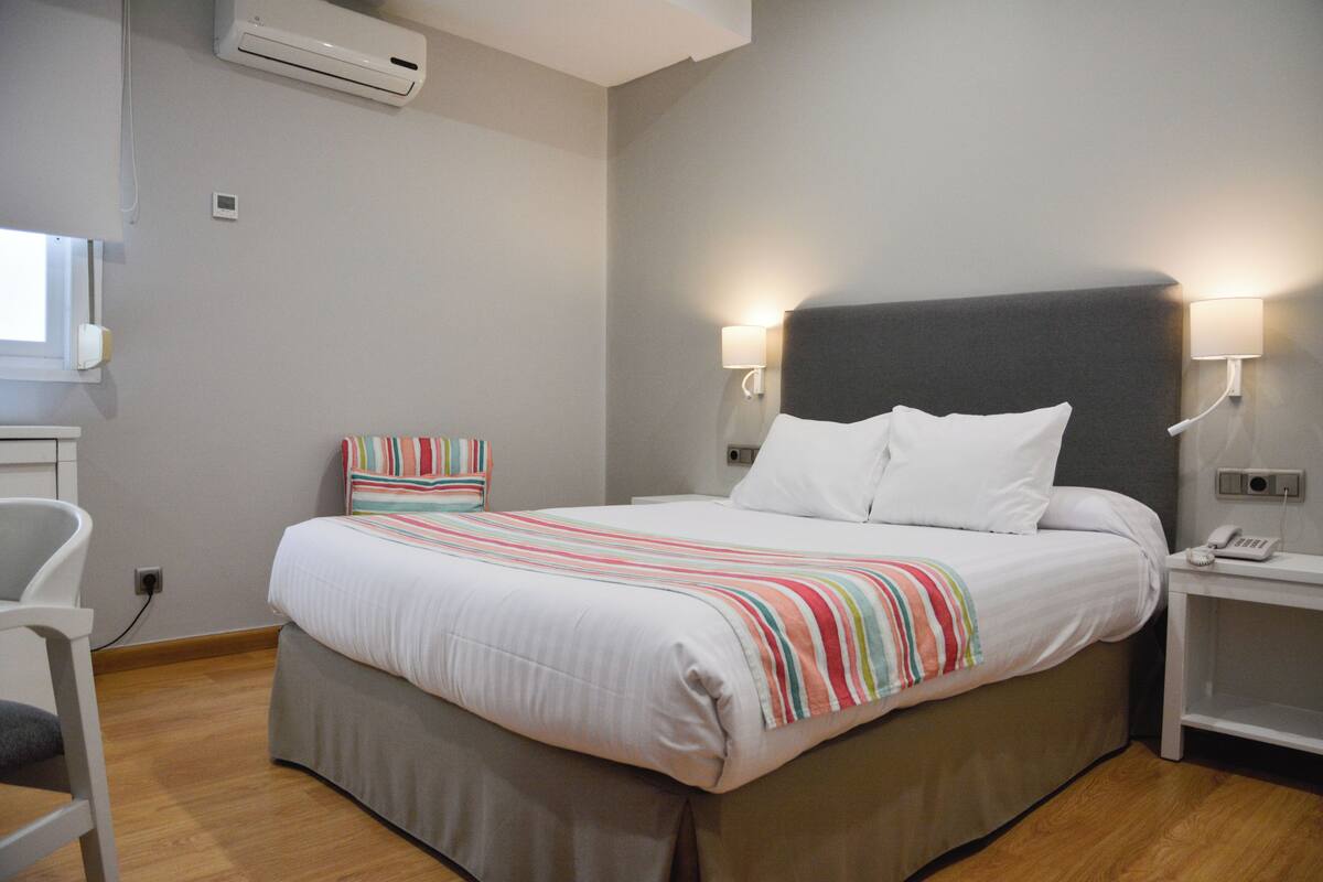Una habitación de hotel con cama, mesitas de noche, una silla y un equipo de aire acondicionado.