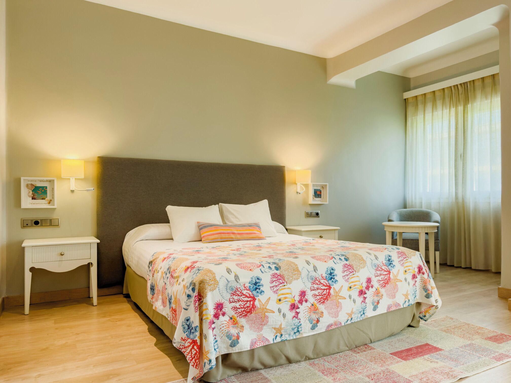 Une chambre d’hôtel comprenant un lit, des tables de chevet, une commode, une lampe, une fenêtre avec des rideaux et une petite table.