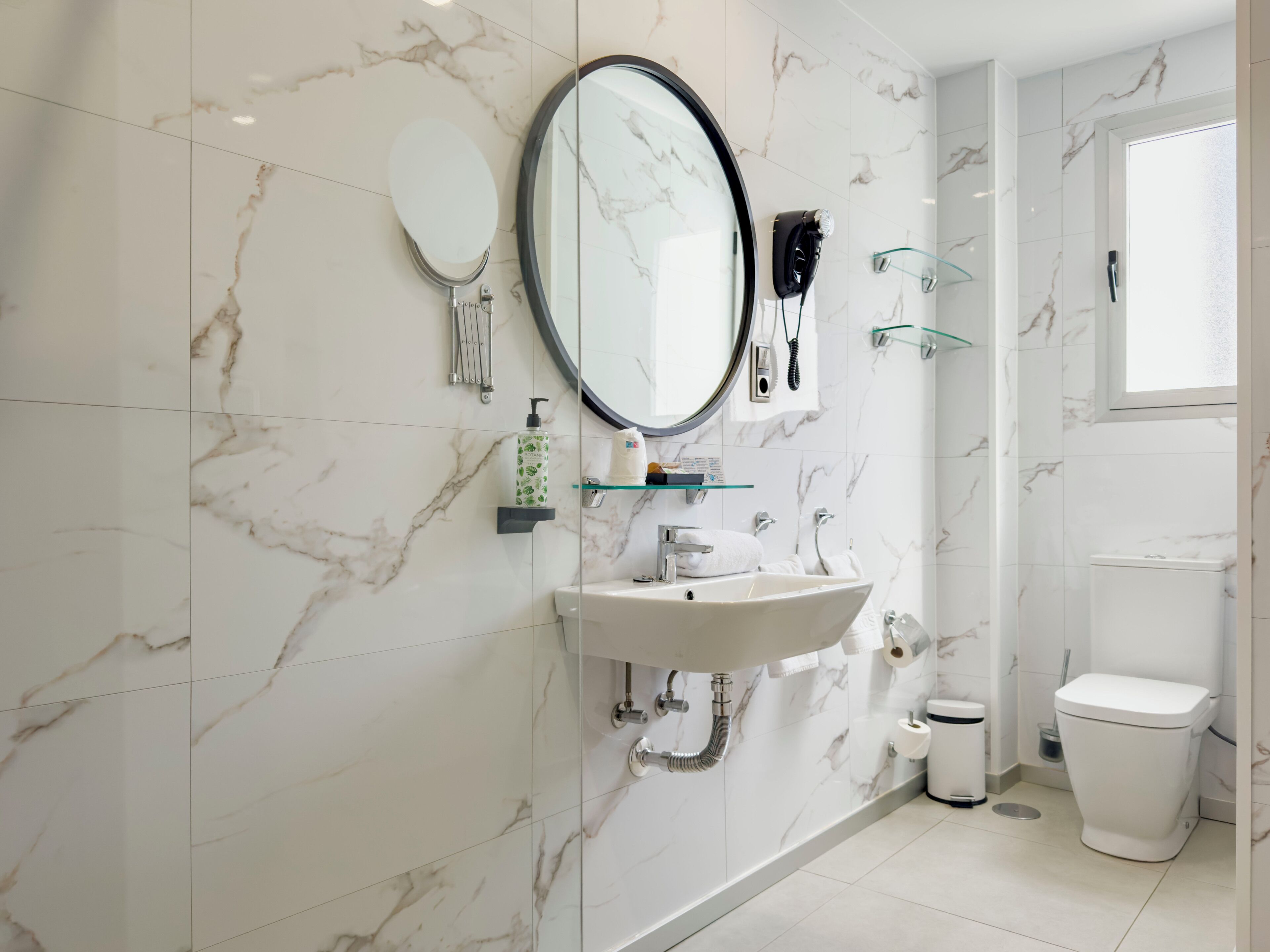 A bathroom with a marble wall, a round mirror, a sink with a faucet, a toilet, and a glass shower enclosure.
