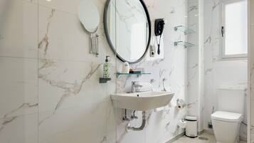 A bathroom with a marble wall, a round mirror, a sink with a faucet, a toilet, and a glass shower enclosure.