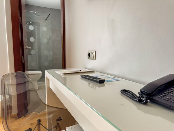 Habitación de hotel con escritorio de vidrio, teléfono, control remoto y ducha visible a través de una puerta de cristal.
