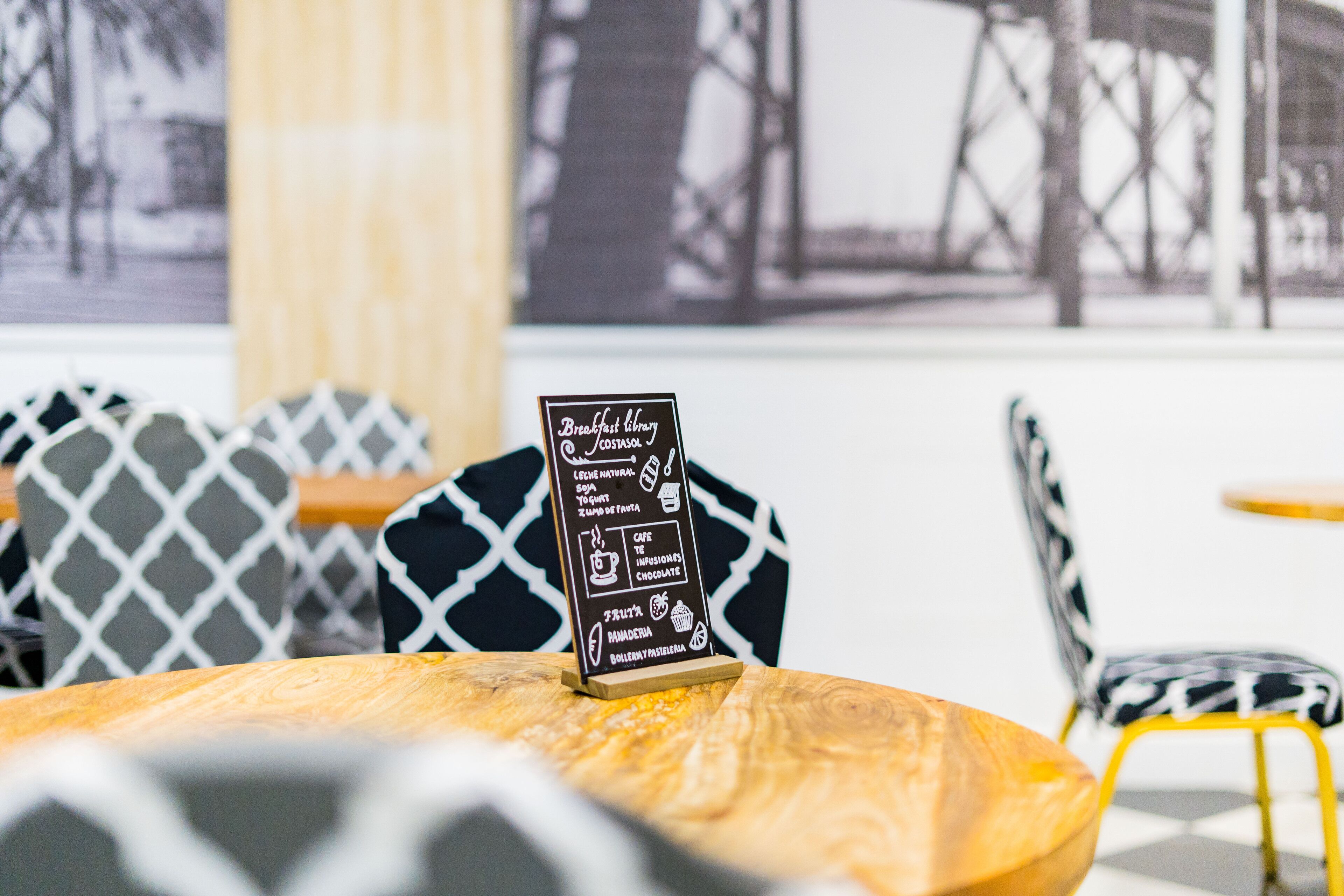 A café with a menu board displaying breakfast items, patterned chairs, and a view of a bridge outside.