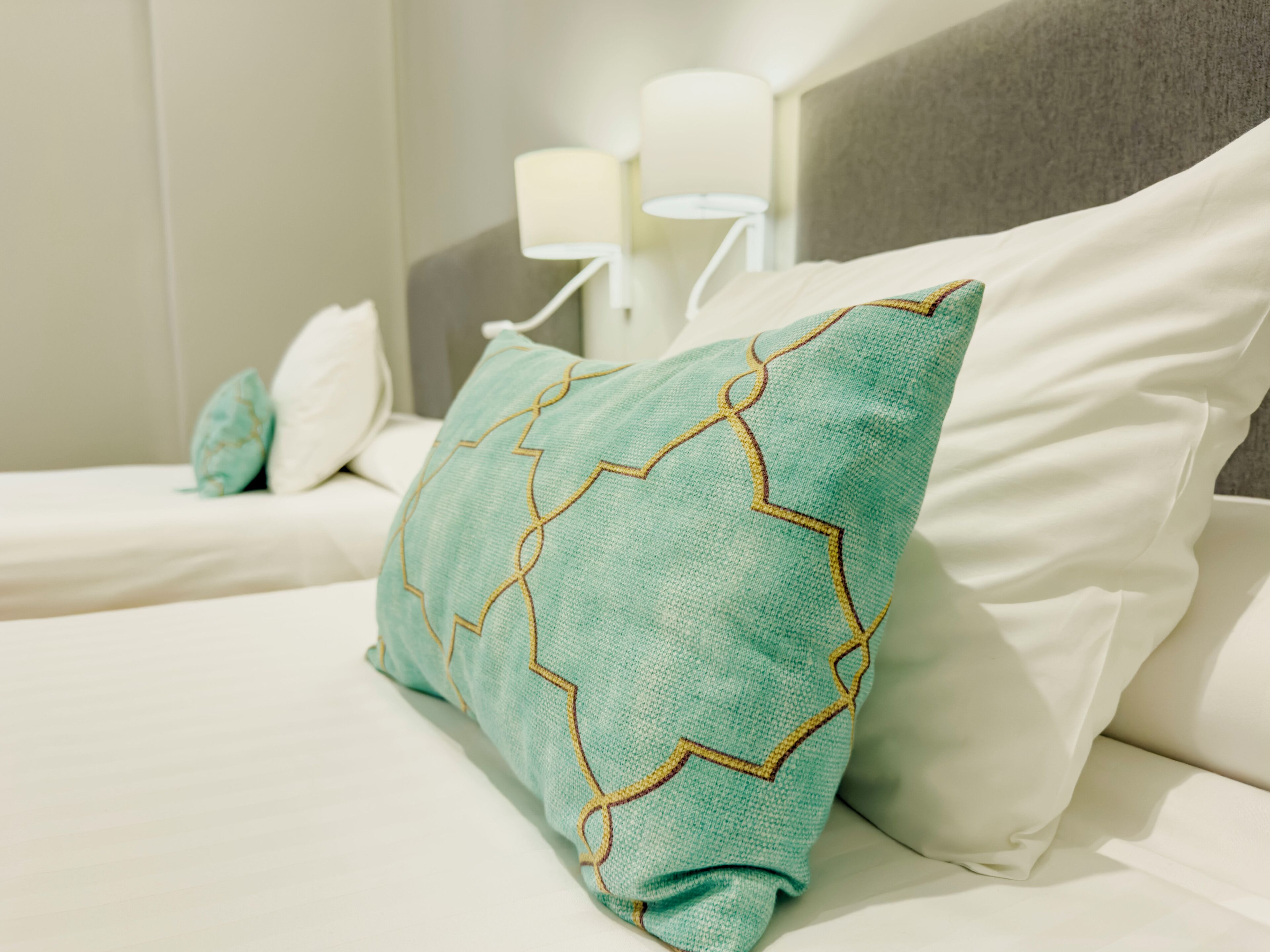 A bed with white bedding and a teal pillow with a geometric pattern.