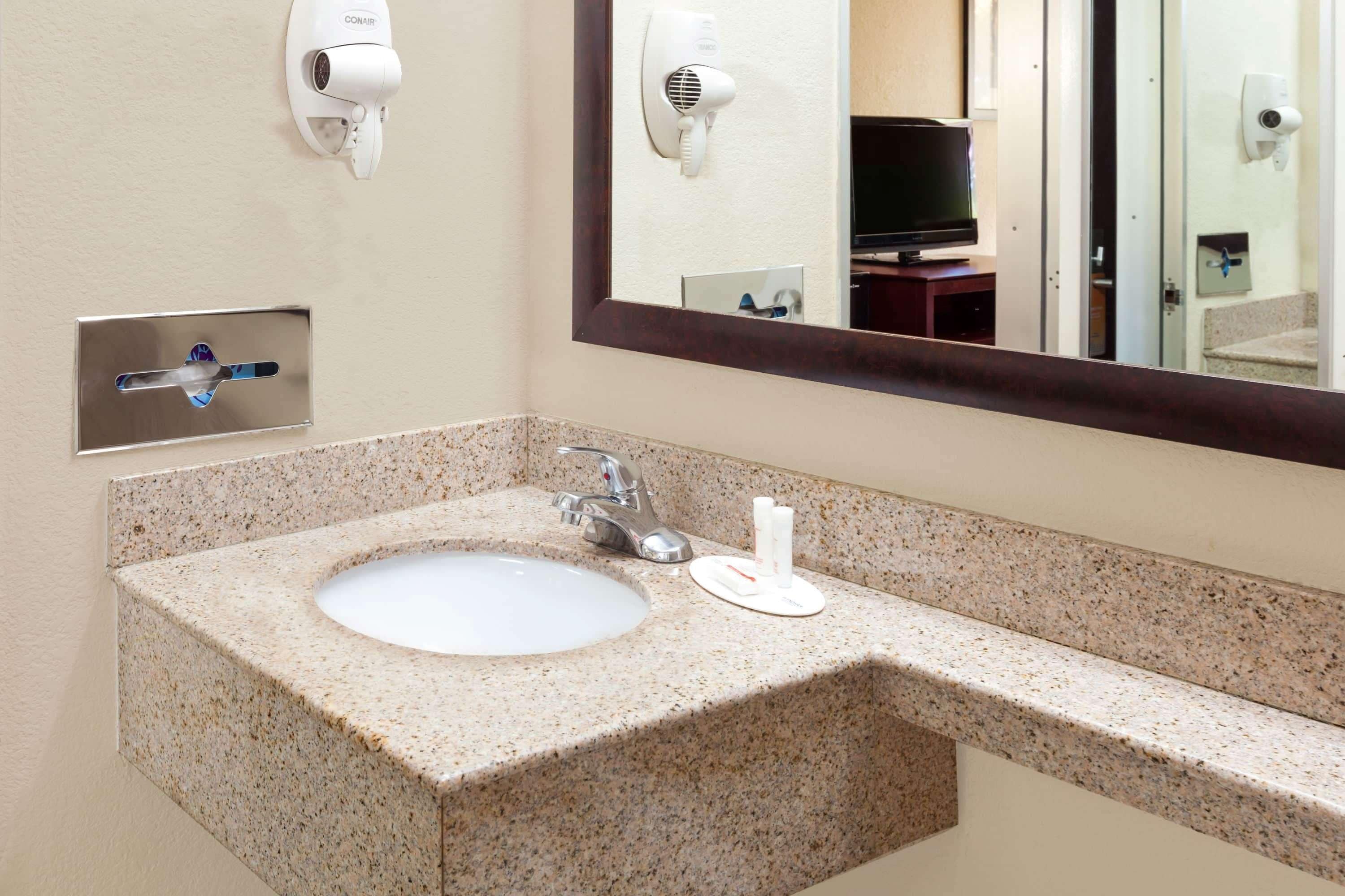 A bathroom with a granite countertop sink, a mirror, a hair dryer, and a television in the background.