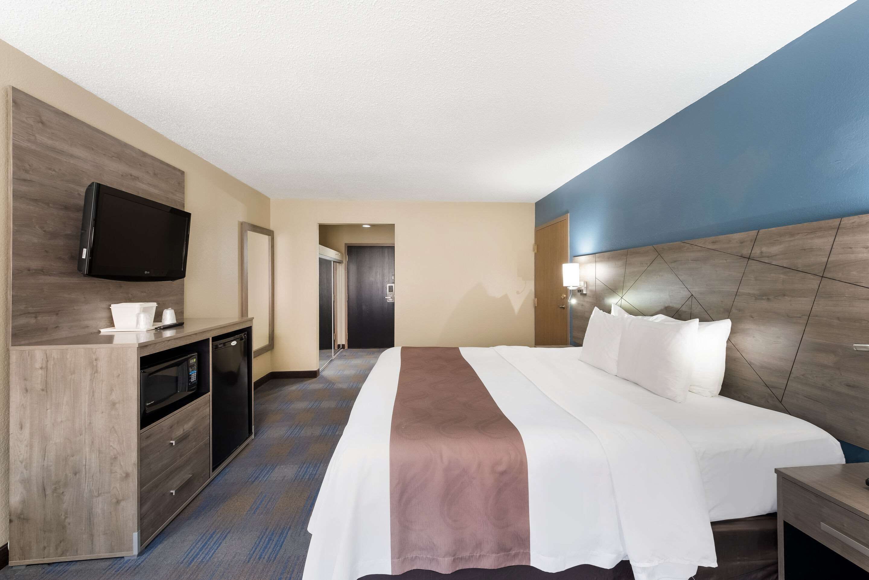 A hotel room with a bed, a TV mounted on the wall, a desk with a microwave, and a door leading to another room.
