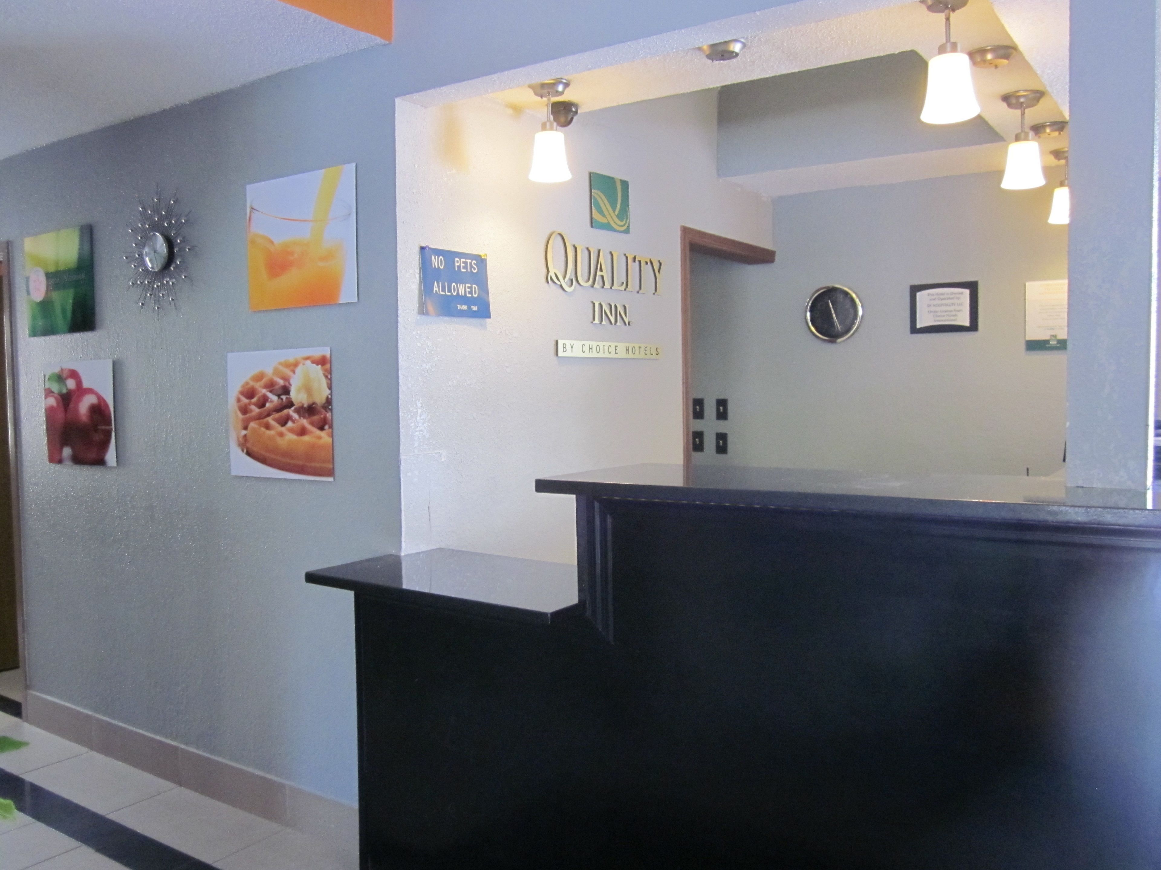 Zona de recepción de un hotel con un mostrador negro, un cartel de 'Quality Inn', un letrero que dice 'No se admiten mascotas' y decoraciones en la pared que incluyen un reloj y fotografías de comida.