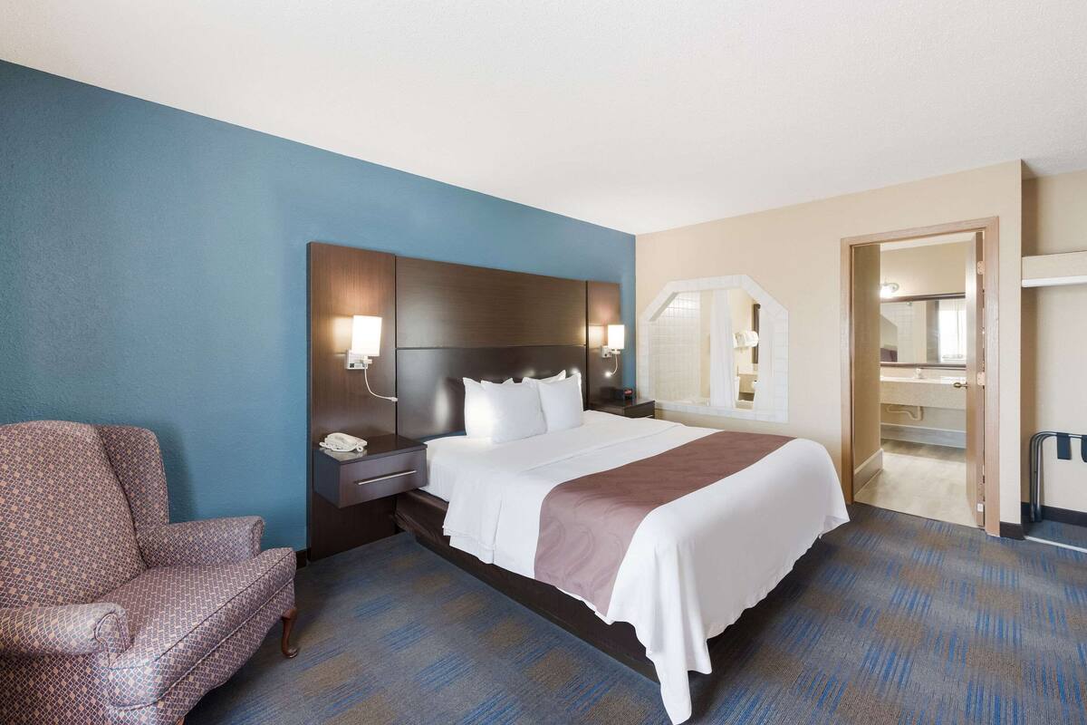 Habitación de hotel con una cama grande, una mesita de noche, una silla y un baño visible a través de una puerta abierta.