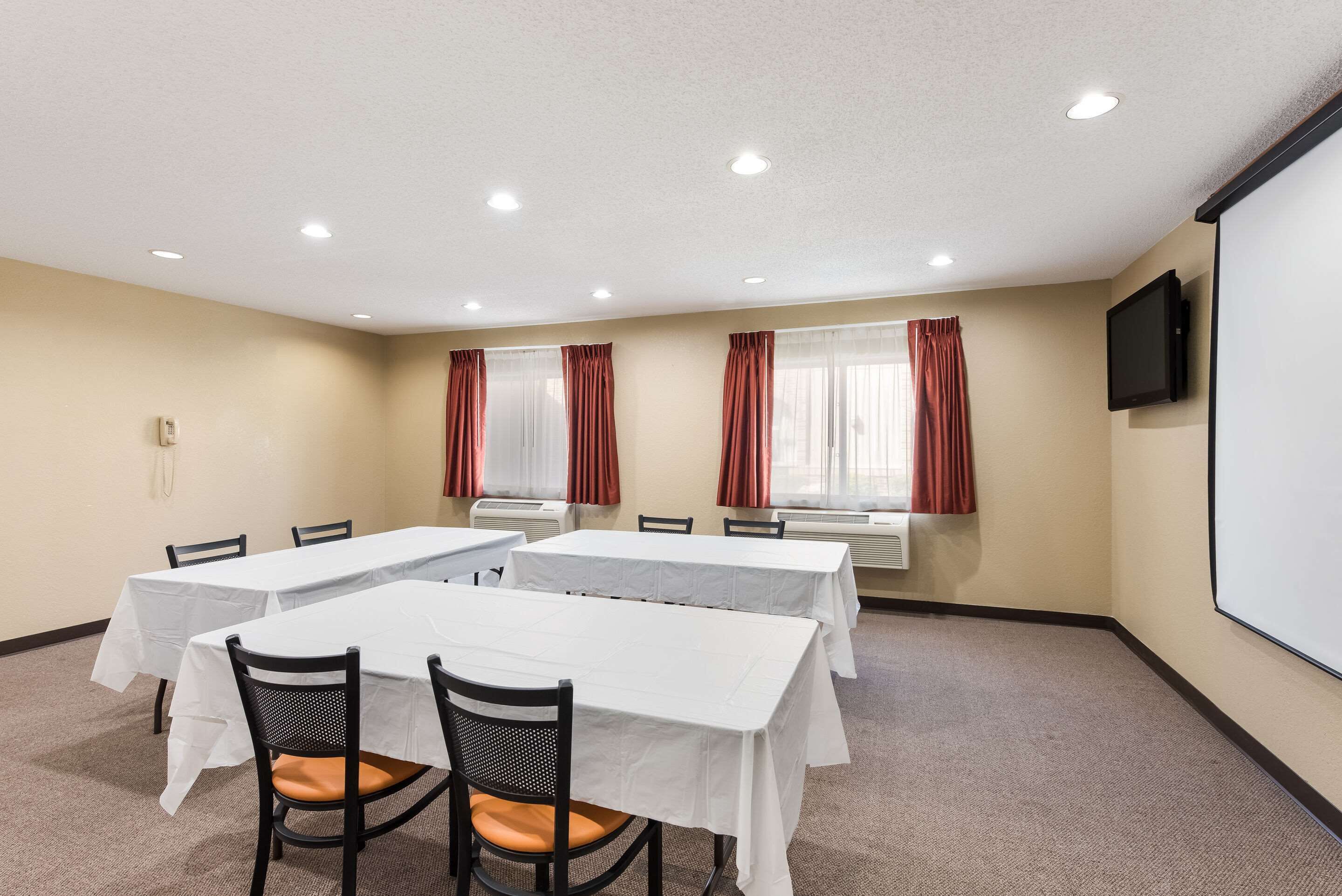 Un salón de conferencias con mesas blancas, sillas negras y cortinas rojas, además de una pantalla de proyector.