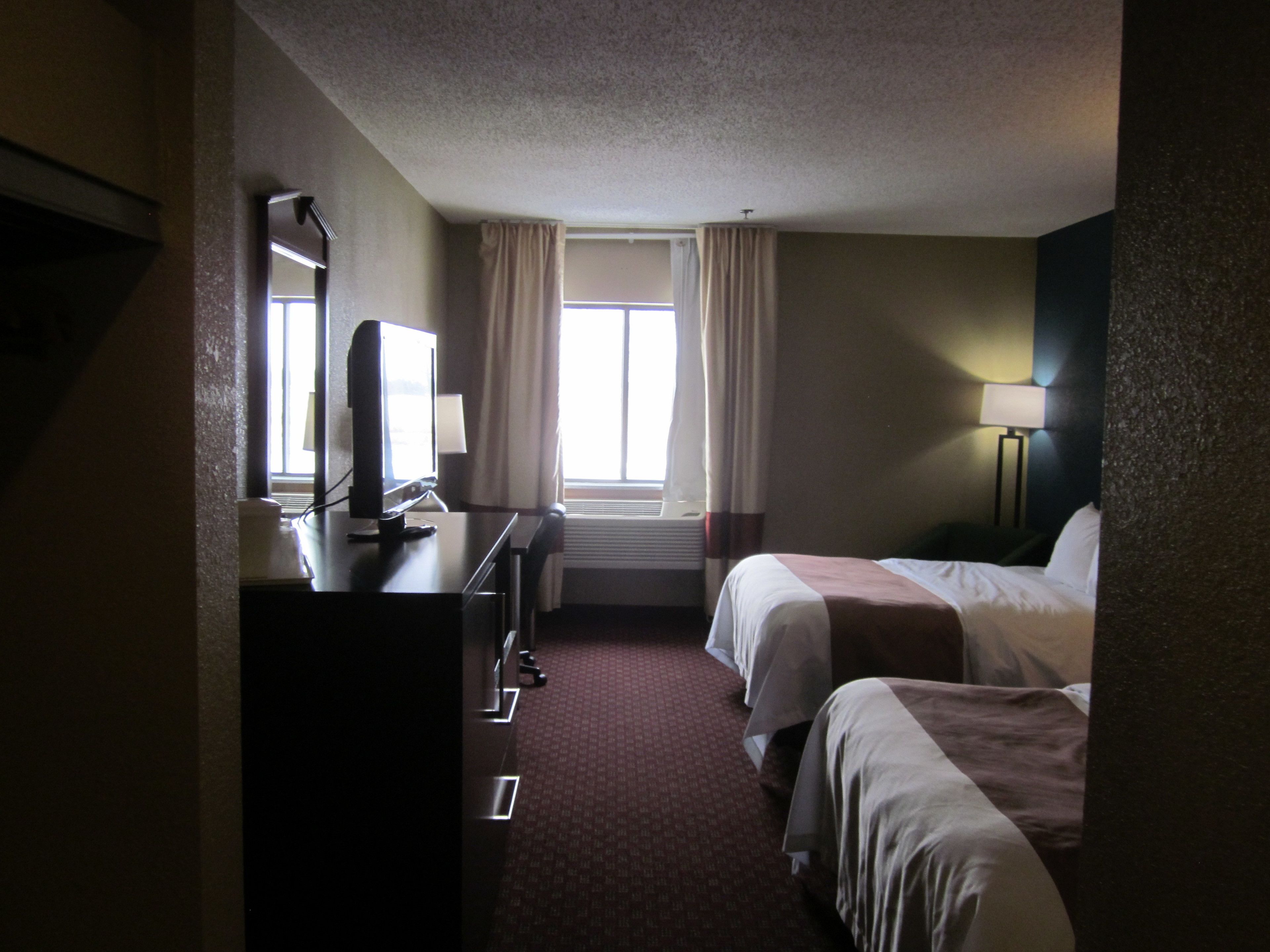 A hotel room with two beds, a desk with a computer, a window with curtains, and a lamp.