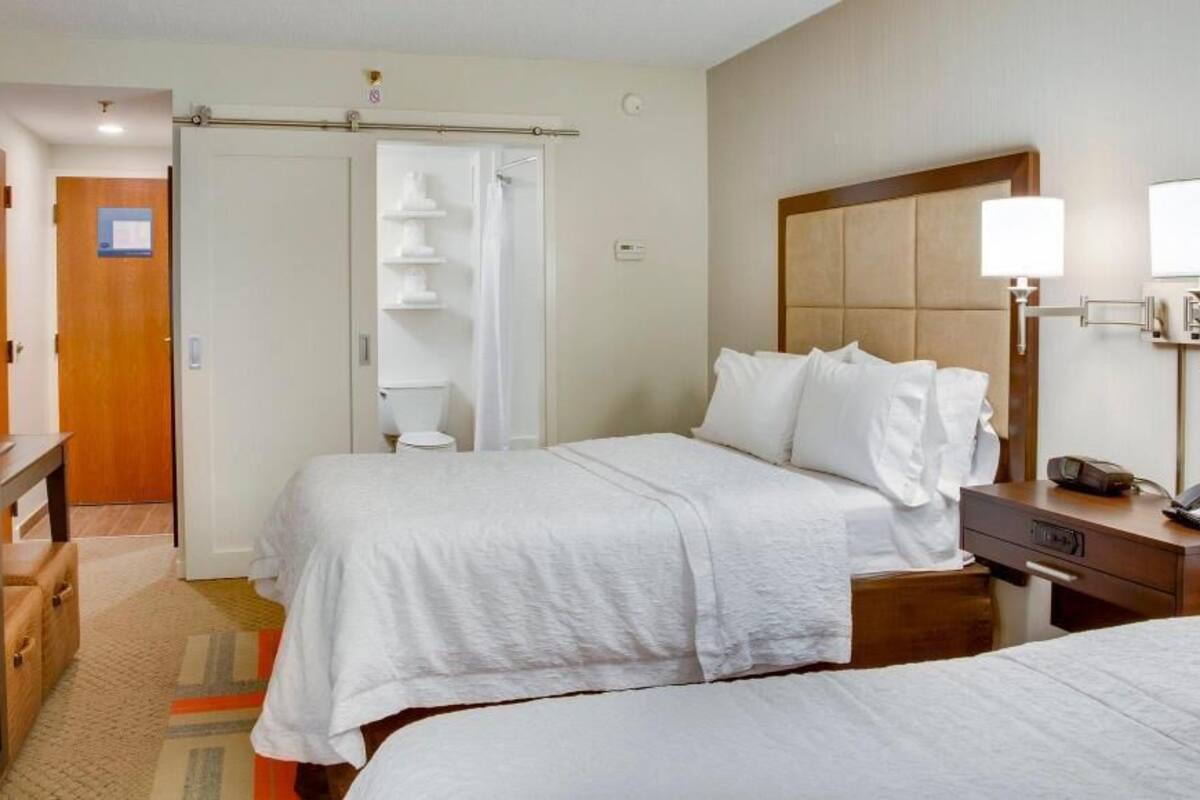 Una habitación de hotel con cama, mesita de noche, lámpara y baño con ducha e inodoro.
