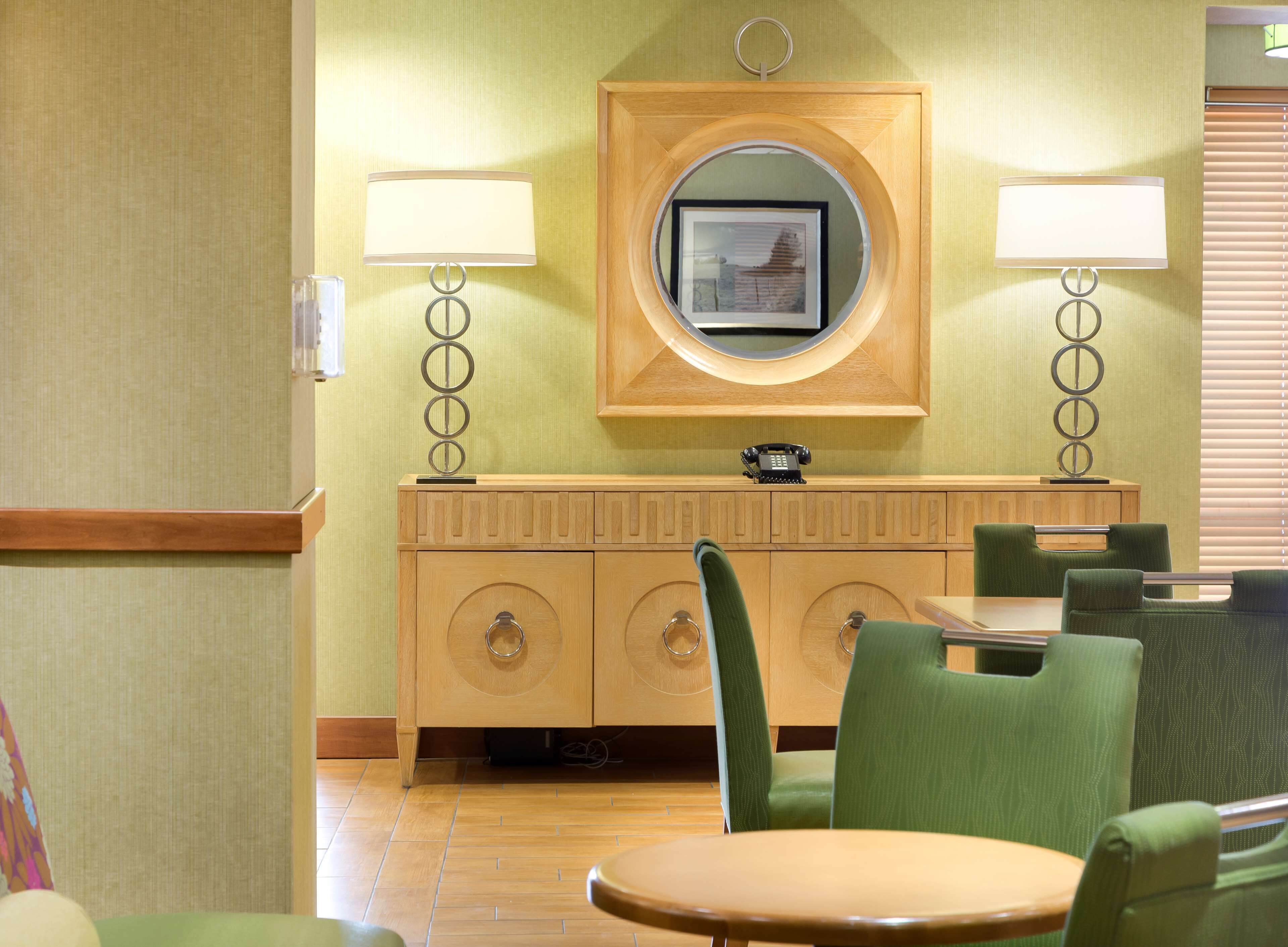 Habitación de hotel con un armario de madera, dos sillas verdes, una mesa redonda y un espejo en la pared.