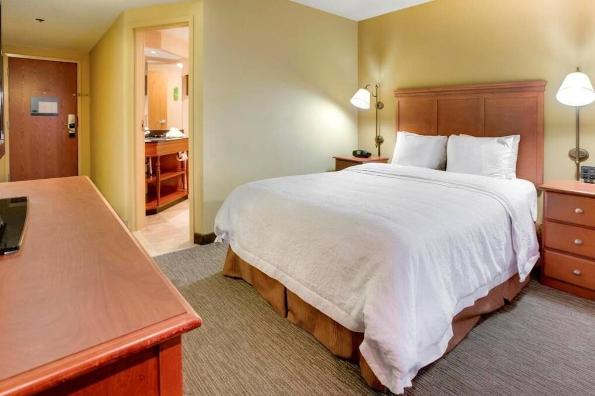 Una habitación de hotel con cama, mesitas de noche, escritorio y baño visible a través de una puerta abierta.