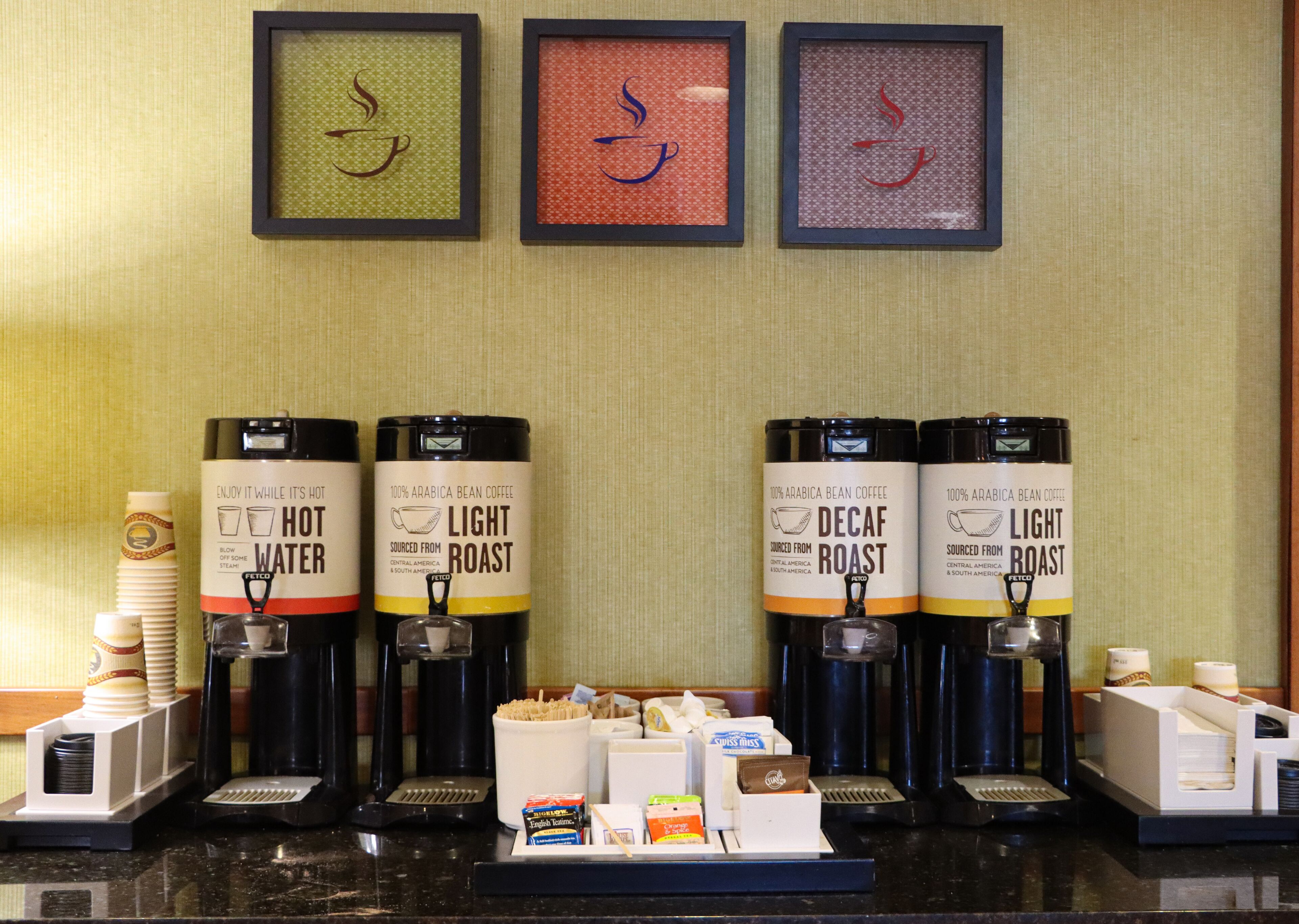 A coffee station with four coffee dispensers labeled 'Hot Water,' 'Light Roast,' 'Decaf Roast,' and 'Light Roast.' There are stacks of disposable cups and a tray with condiments and napkins.
