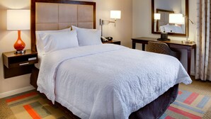 Ein Hotelzimmer mit einem großen Bett, einem Schreibtisch, einem Stuhl, einem Nachttisch mit Lampe, einem Telefon und einem Spiegel.