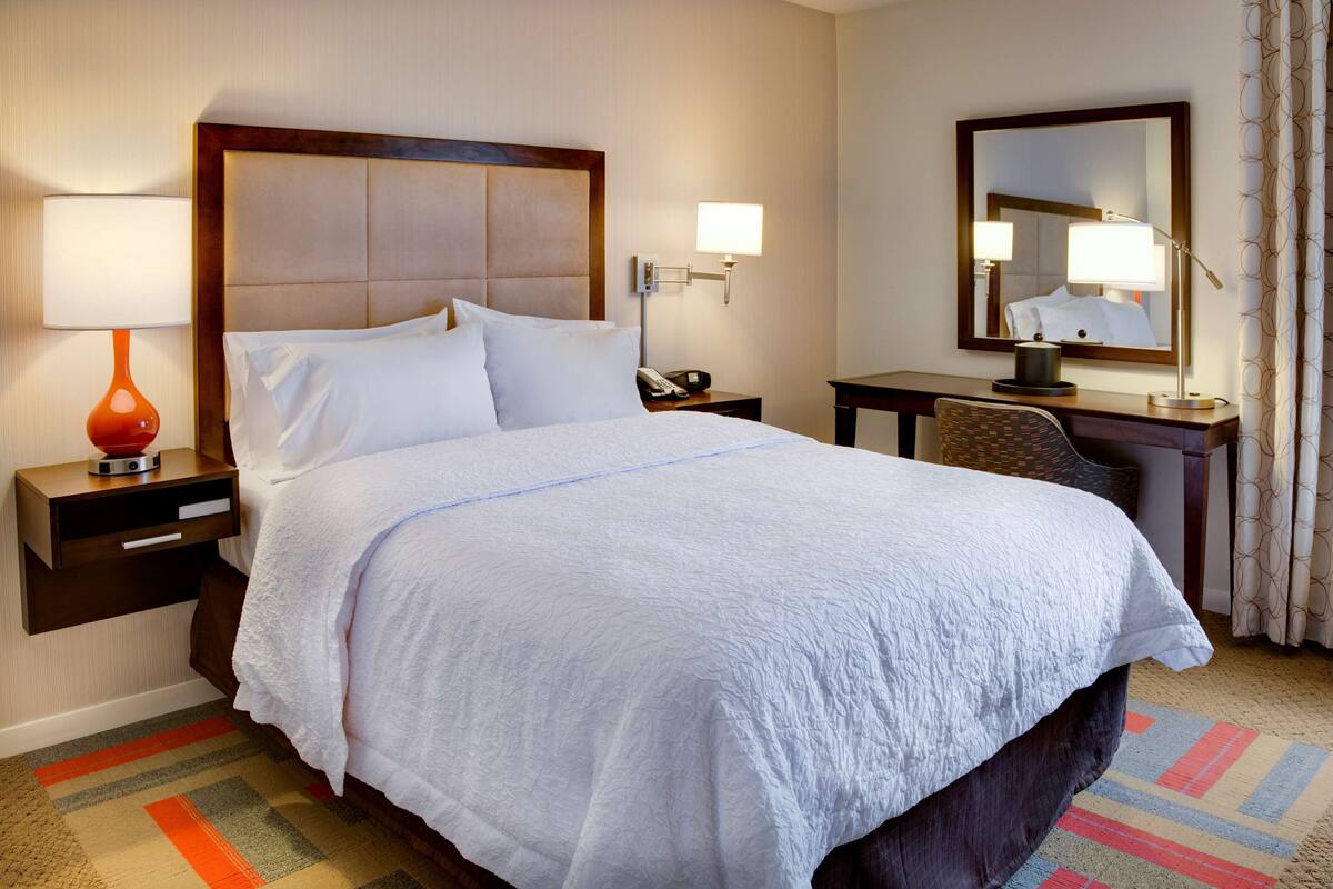 Habitación de hotel con una cama grande, un escritorio, una silla, una mesita de noche con lámpara, teléfono y espejo.